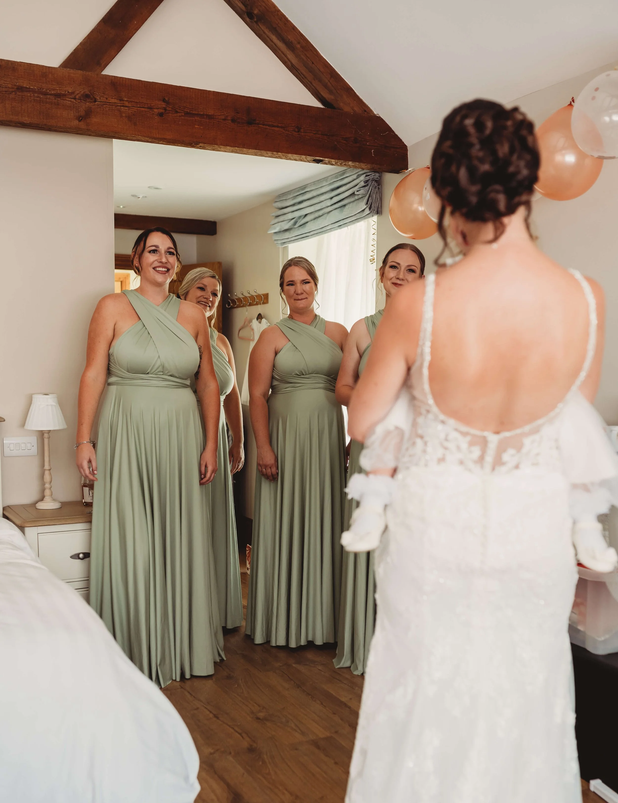 Bride in white lace wedding dress facing four bridesmaids in light green dresses, smiling and looking at her, in a cozy bedroom with wood beams and soft natural light.