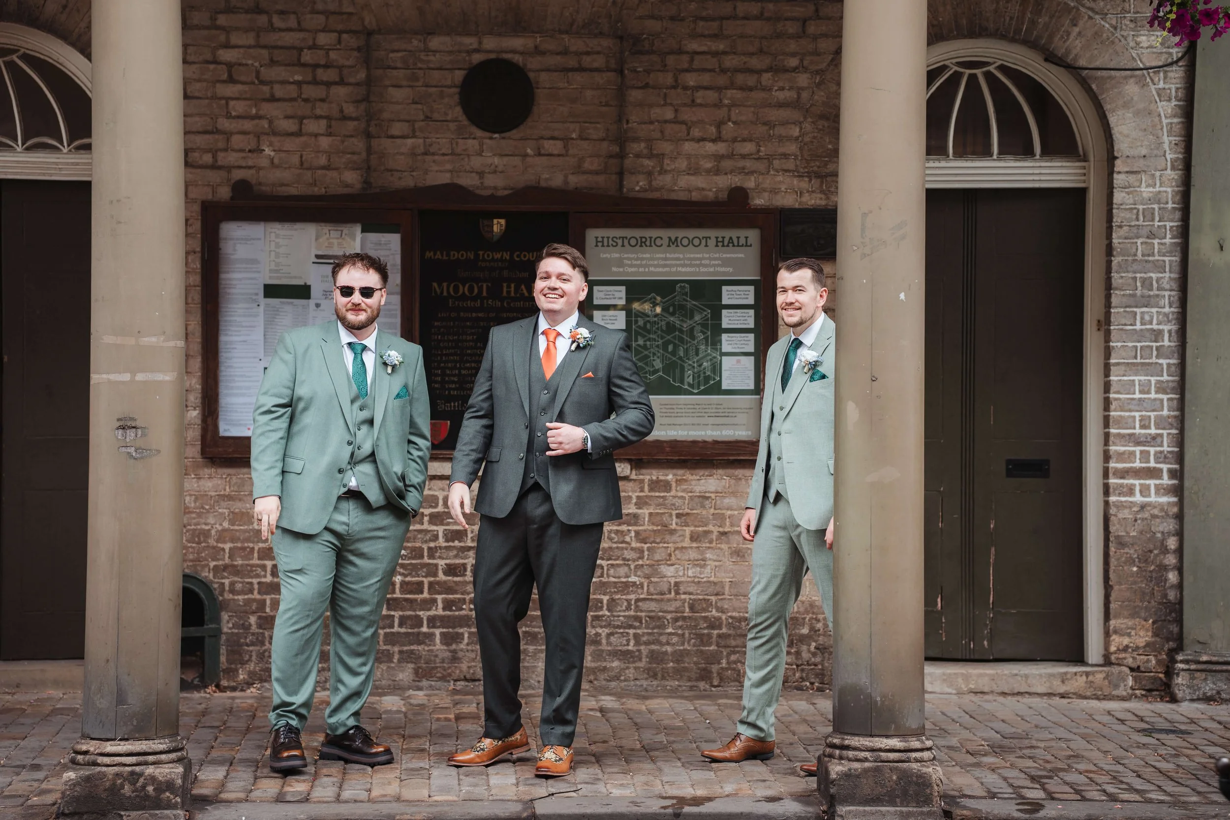 Three men in suits standing on a cobblestone street in front of a brick wall, with two large columns framing the image and a wooden sign behind them that reads 'HISTORIC MOOT HALL.' All are dressed in formal attire with boutonnières, smiling and posi