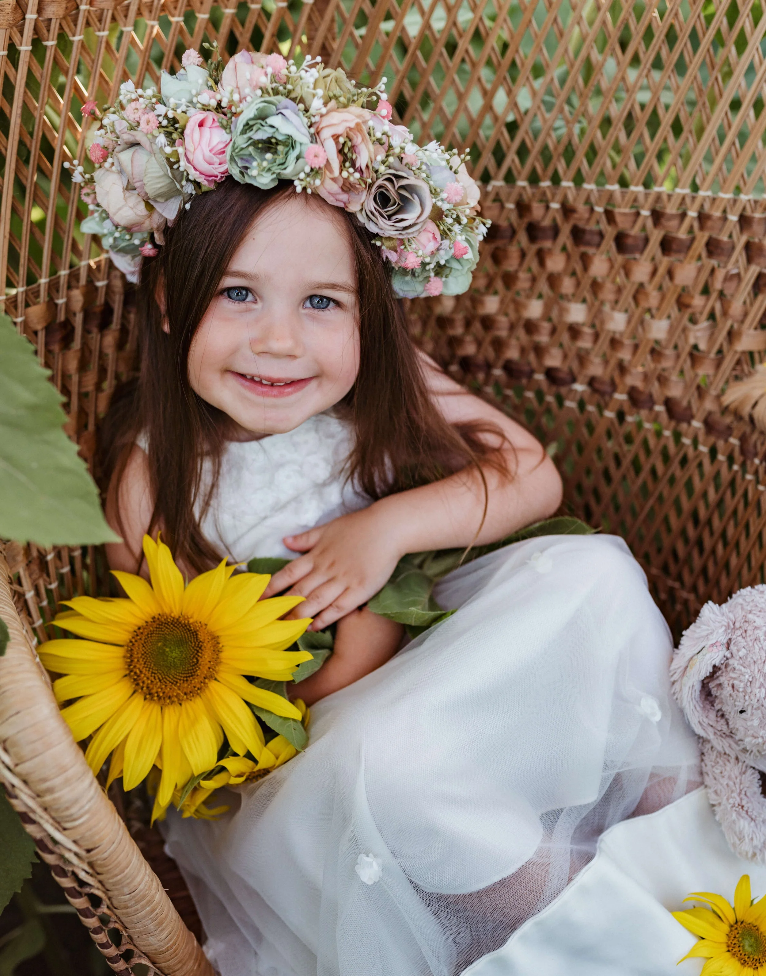 A young girl with long brown hair and blue eyes, wearing a floral crown, sitting in a wicker chair surrounded by flowers, with a sunflower in her lap and smiling at the camera.
