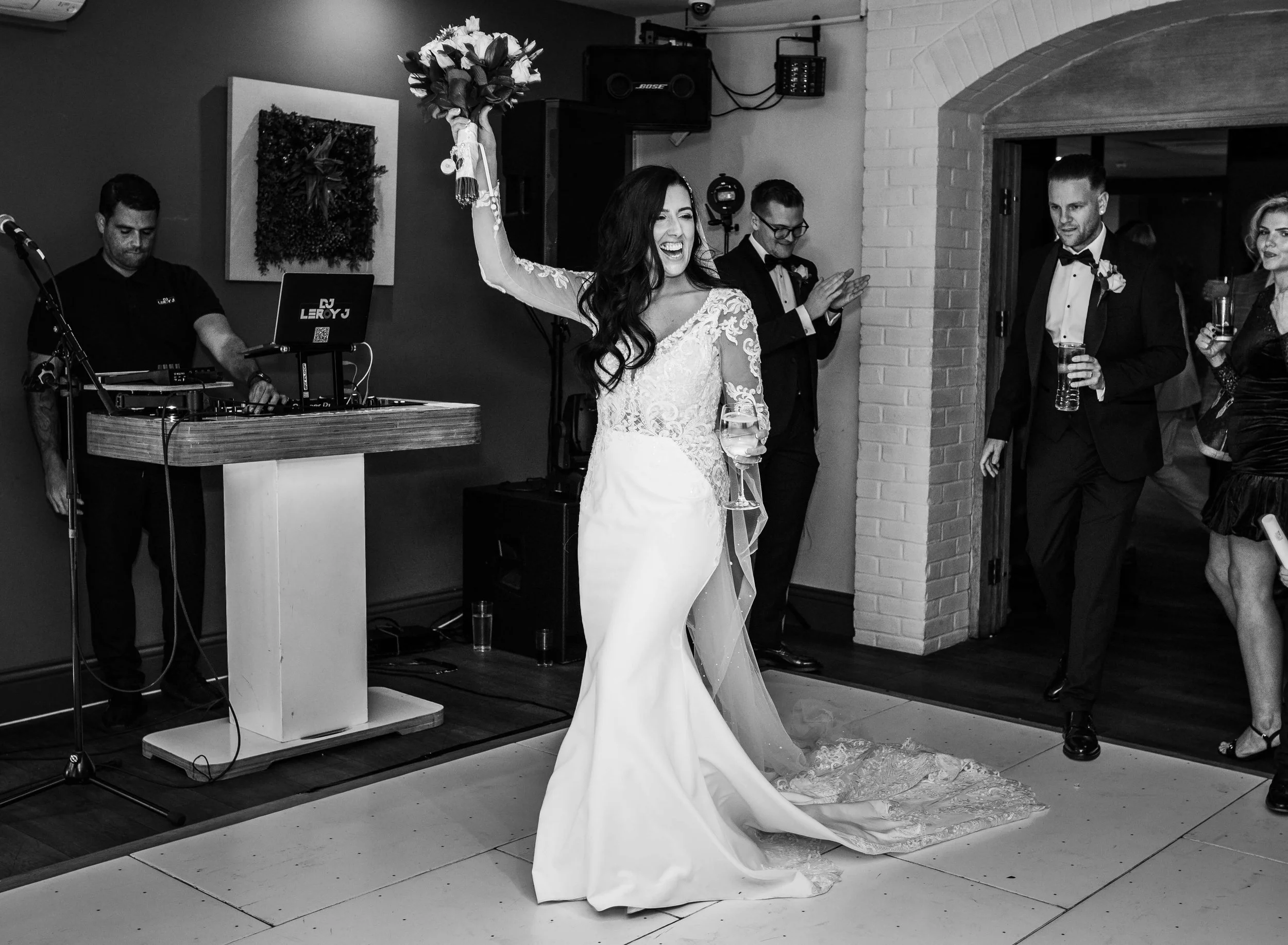 Black and white photo of a bride at her wedding reception, holding a bouquet above her head and smiling, with guests clapping and drinking beer in a decorated indoor venue.
