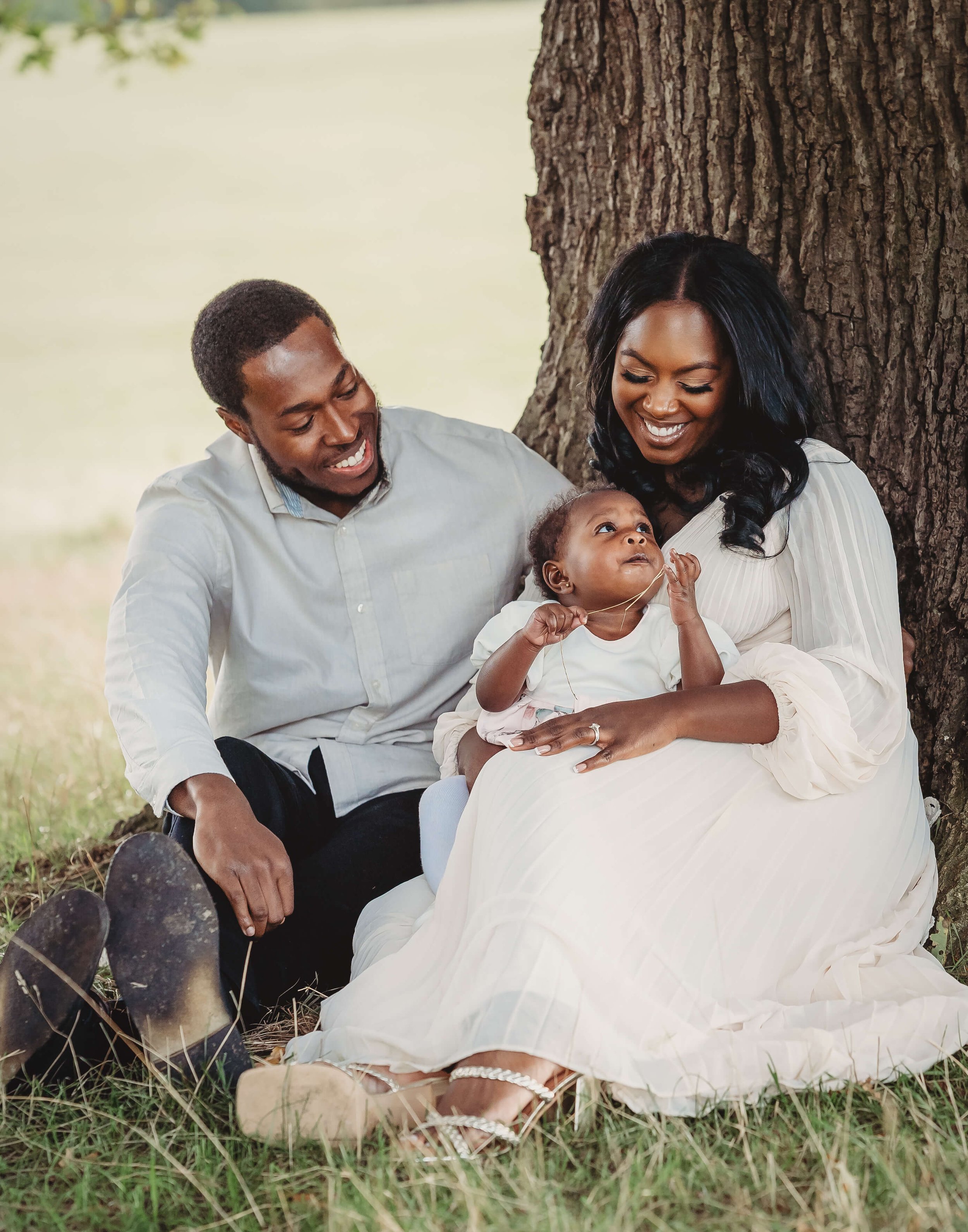 A happy family of three sitting under a tree on grass. The woman is holding a young girl, and the man is sitting beside them. They are smiling and enjoying a moment together outdoors.