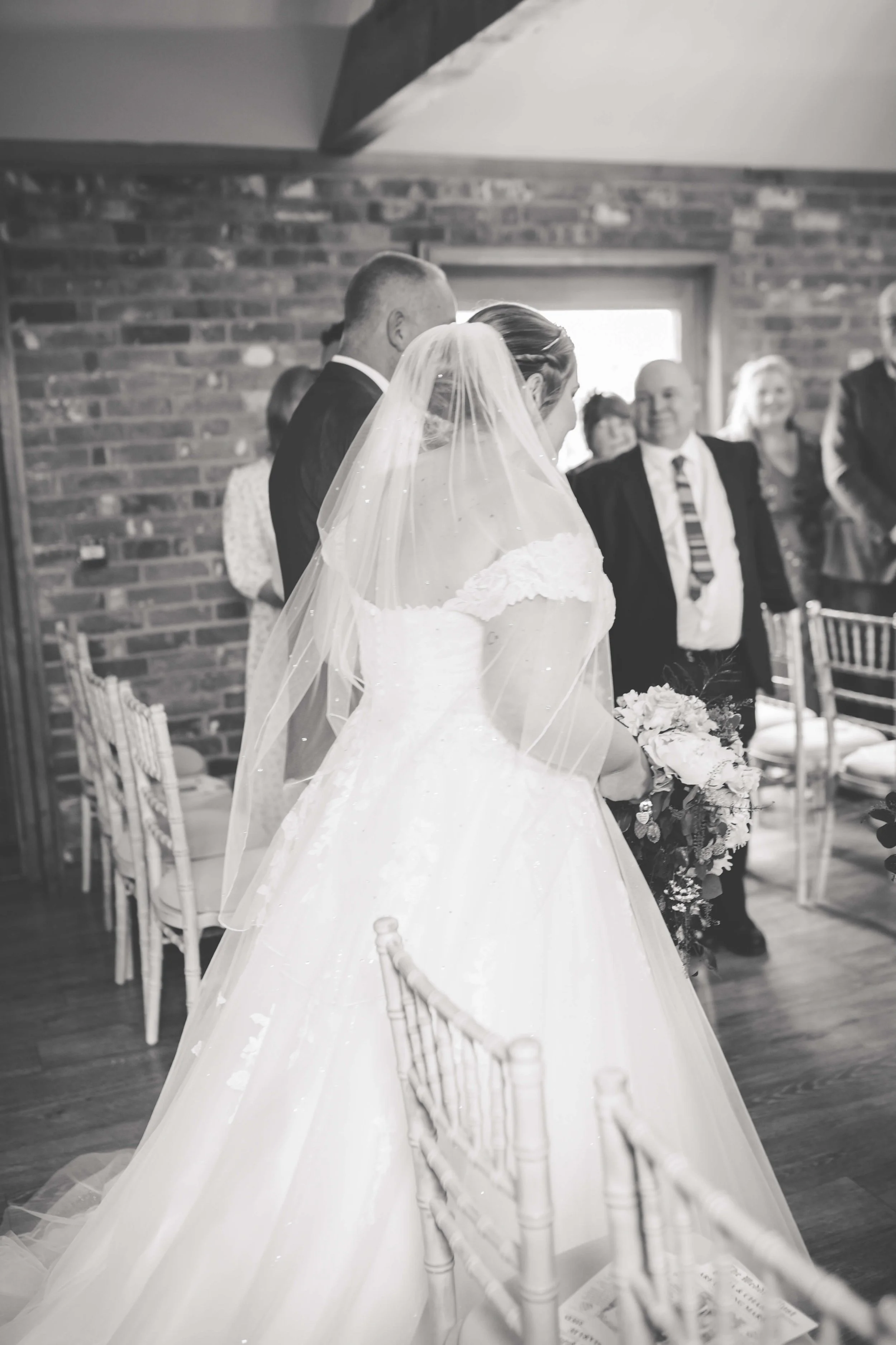 A bride in a wedding dress and veil holding a bouquet, walking down the aisle with a groom in a tuxedo, during a wedding ceremony in a rustic brick room, with guests watching.