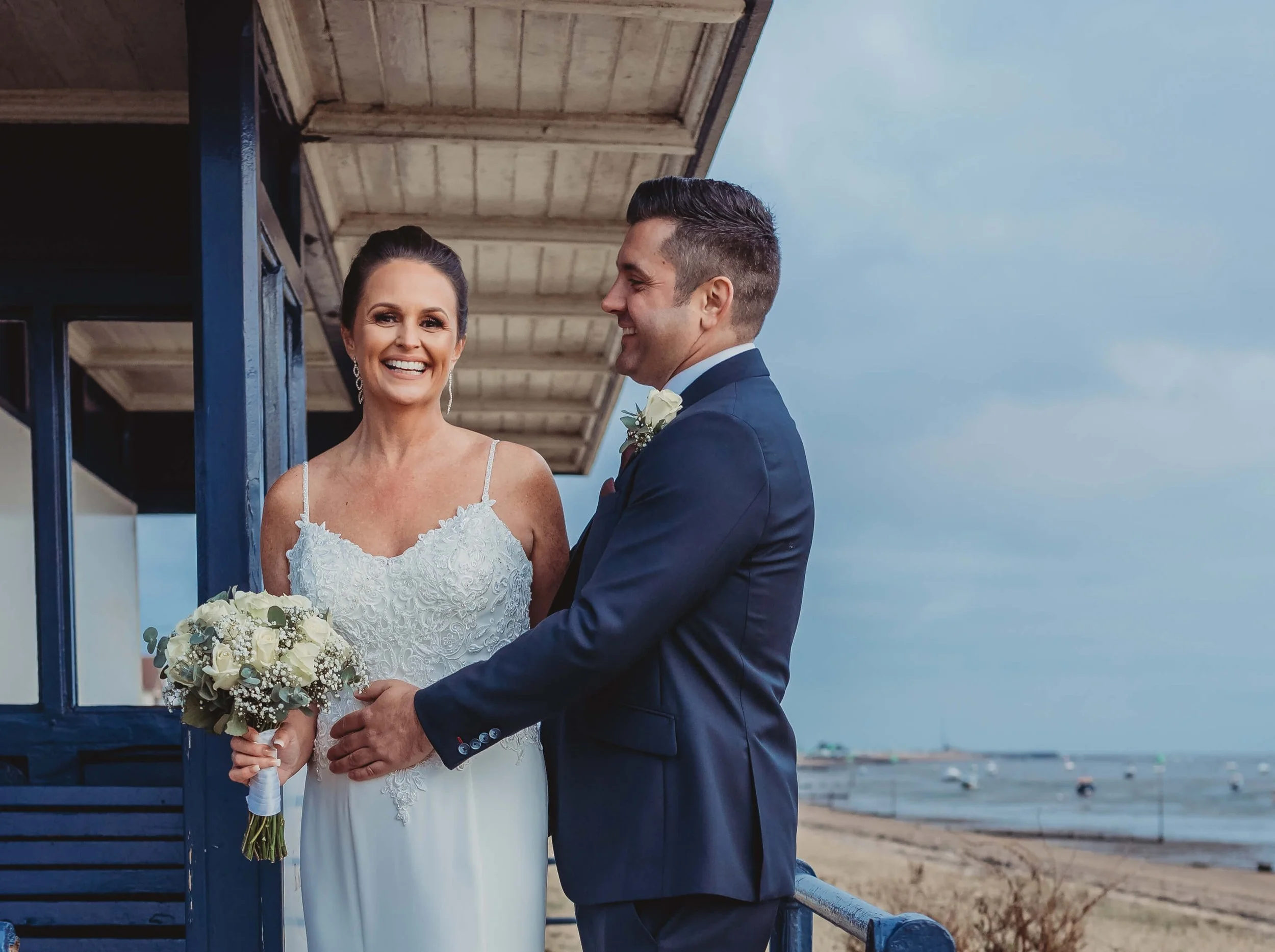 A bride and groom on a beach pier, smiling at each other, with the bride holding a bouquet of white flowers and the groom in a navy suit, overlooking the ocean with boats and a cloudy sky in the background.