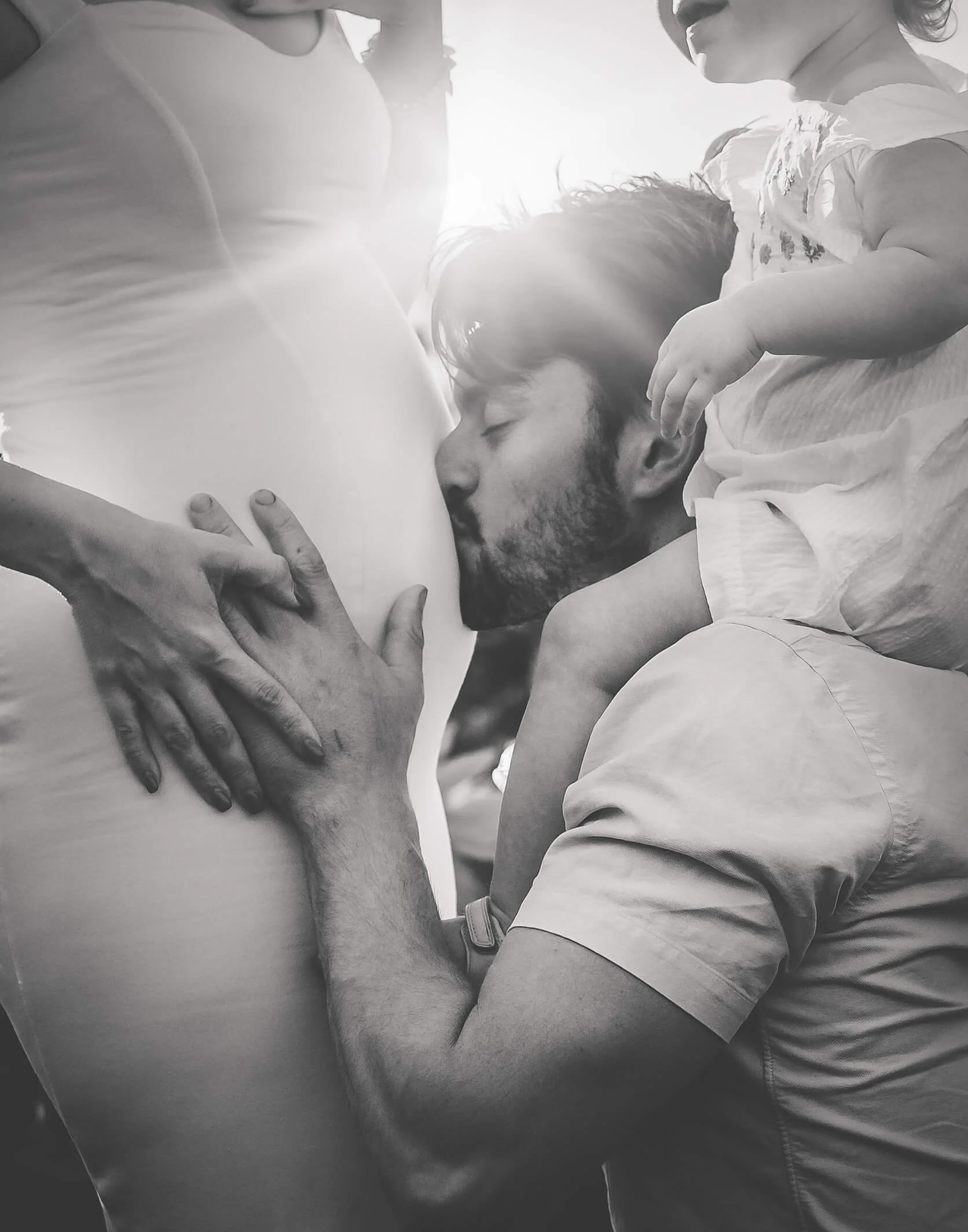 Man kissing a pregnant woman's belly while her hand rests on her stomach, and two young children stand nearby, all in black and white.