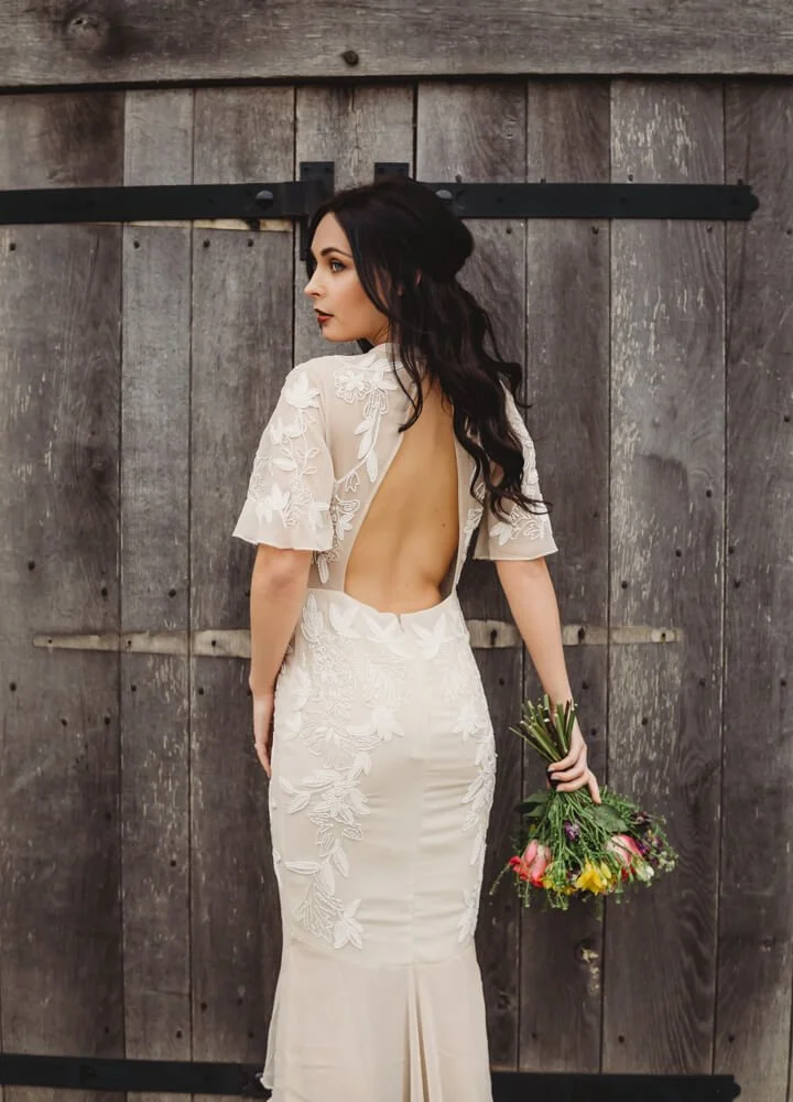 A woman with dark hair in a white lace backless dress holding a bouquet of colorful flowers standing in front of a rustic wooden door.