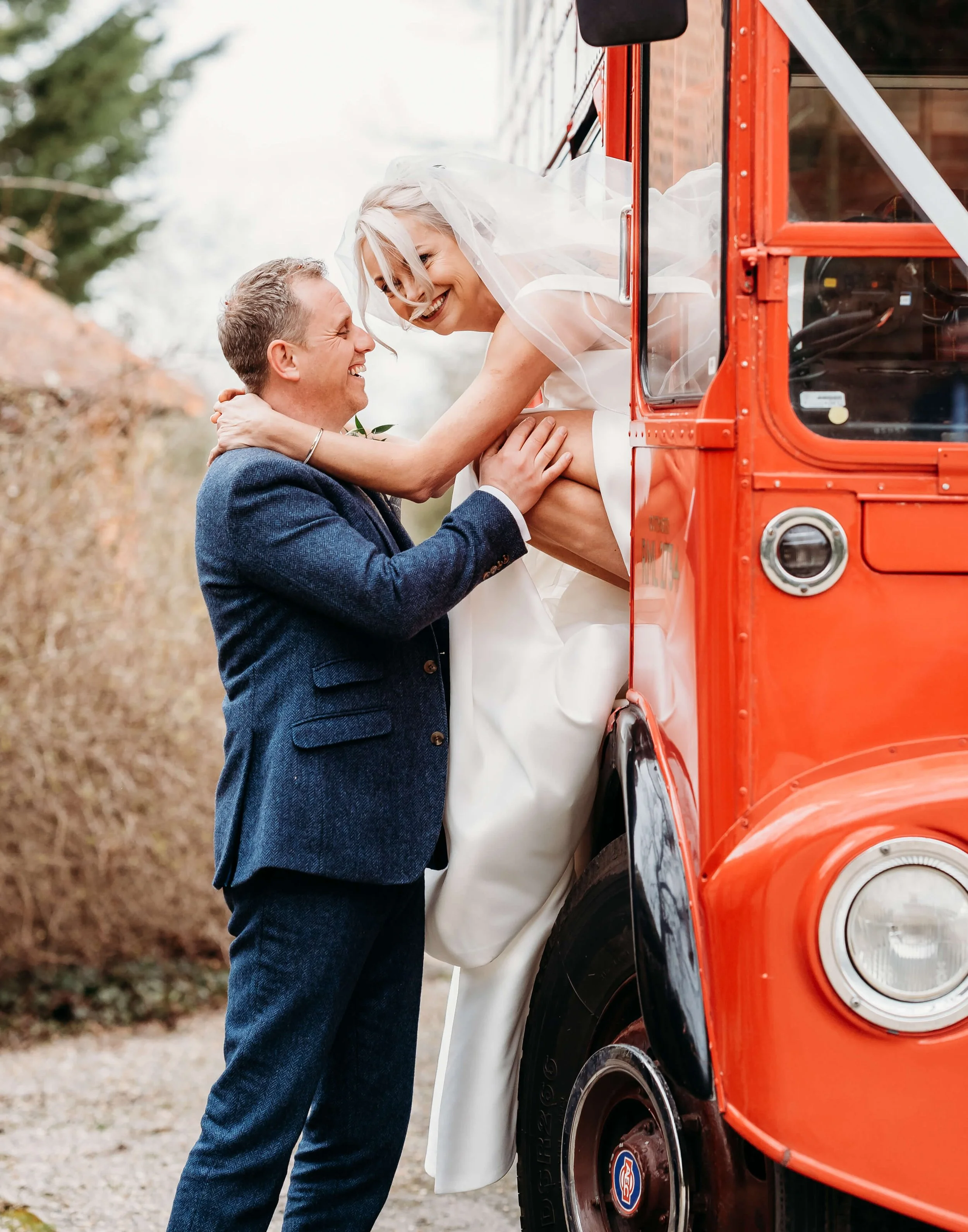 A joyful bride in a white wedding dress and veil sits on a red vehicle while a groom in a navy suit lifts her up, smiling at each other outdoors.