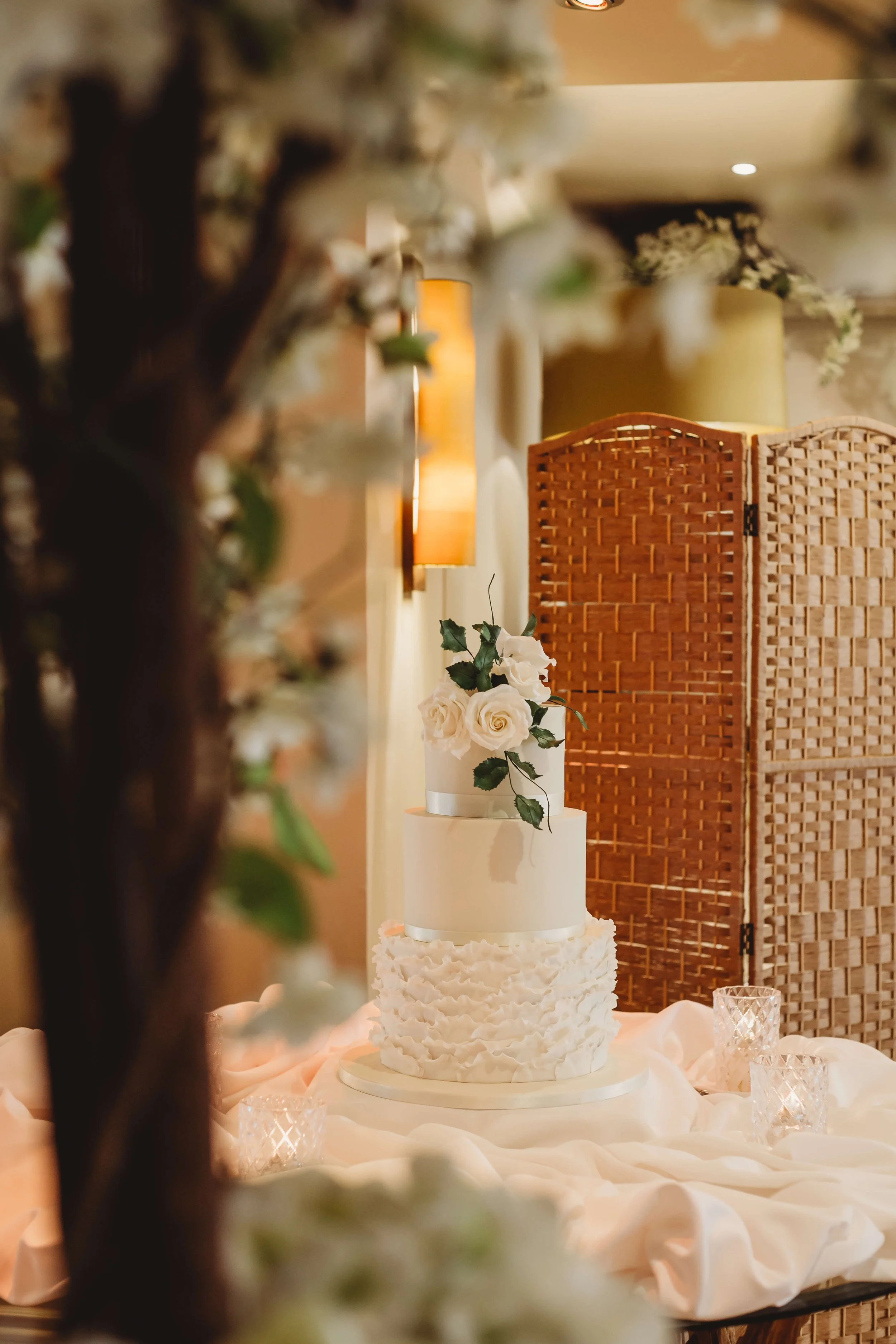 A wedding cake with three tiers, decorated with white roses and green leaves, surrounded by a pink fabric and small glass candle holders.