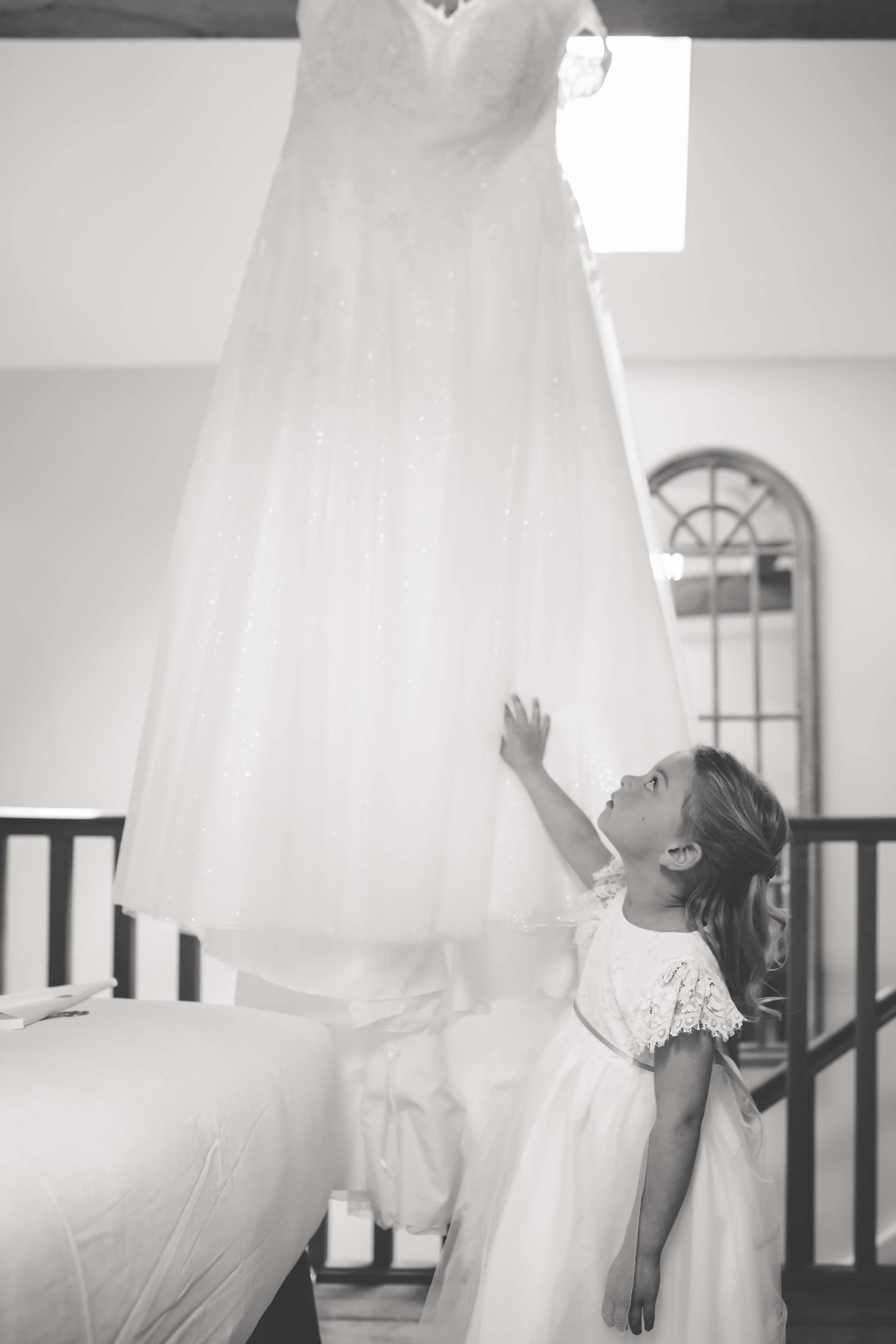 A young girl looking up at a wedding dress hanging above her in a bright room.