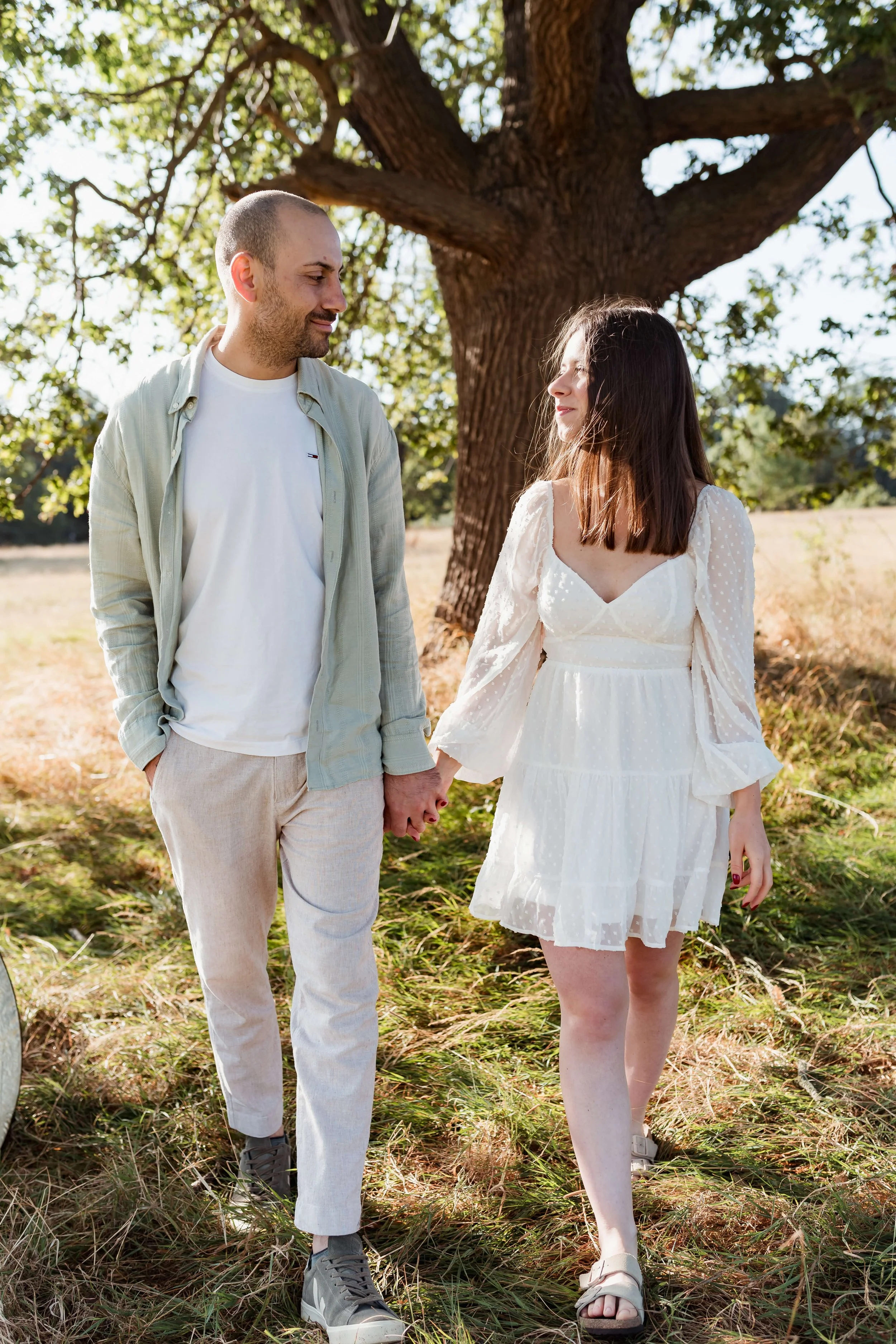 A couple holding hands during an outdoor photoshoot, standing under a large tree, with sunlight filtering through the leaves.