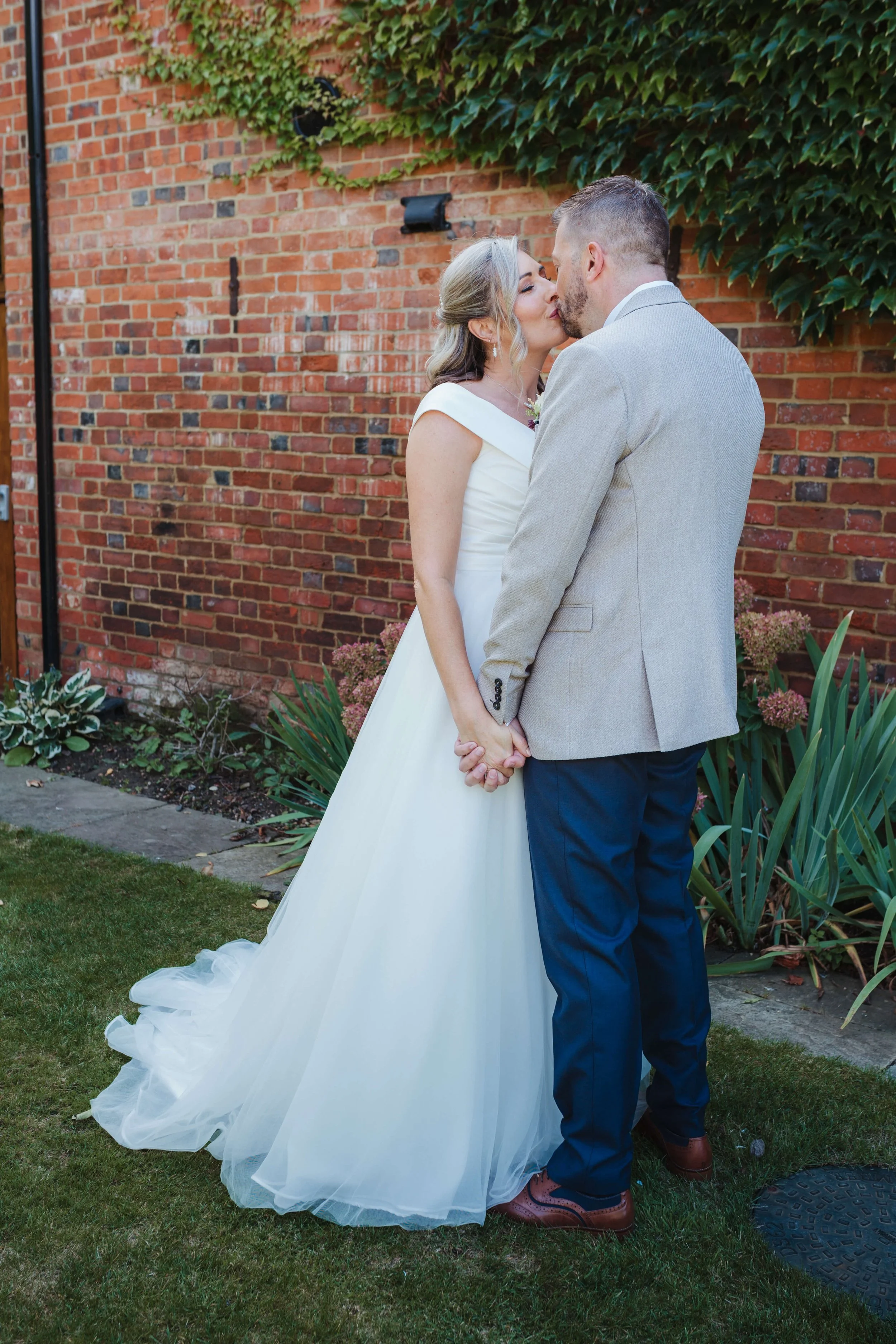 A bride and groom standing close, holding hands, sharing a kiss outdoors against a brick wall with ivy, surrounded by green plants.