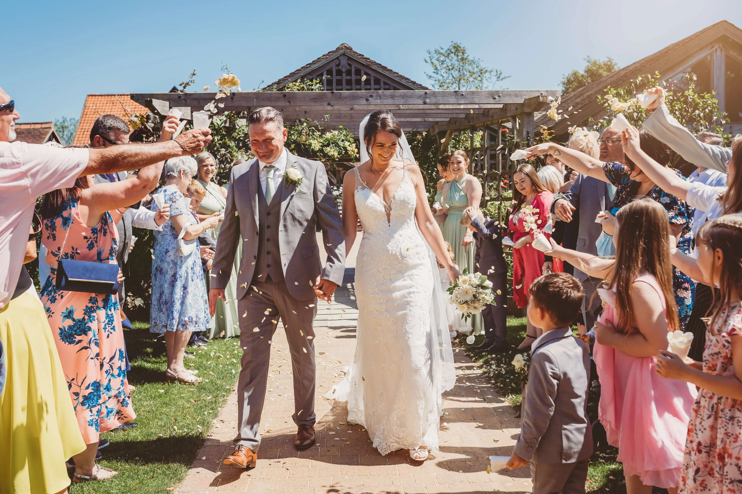 Bride and groom walking hand-in-hand through a crowd of wedding guests, surrounded by confetti, outdoors on a sunny day.