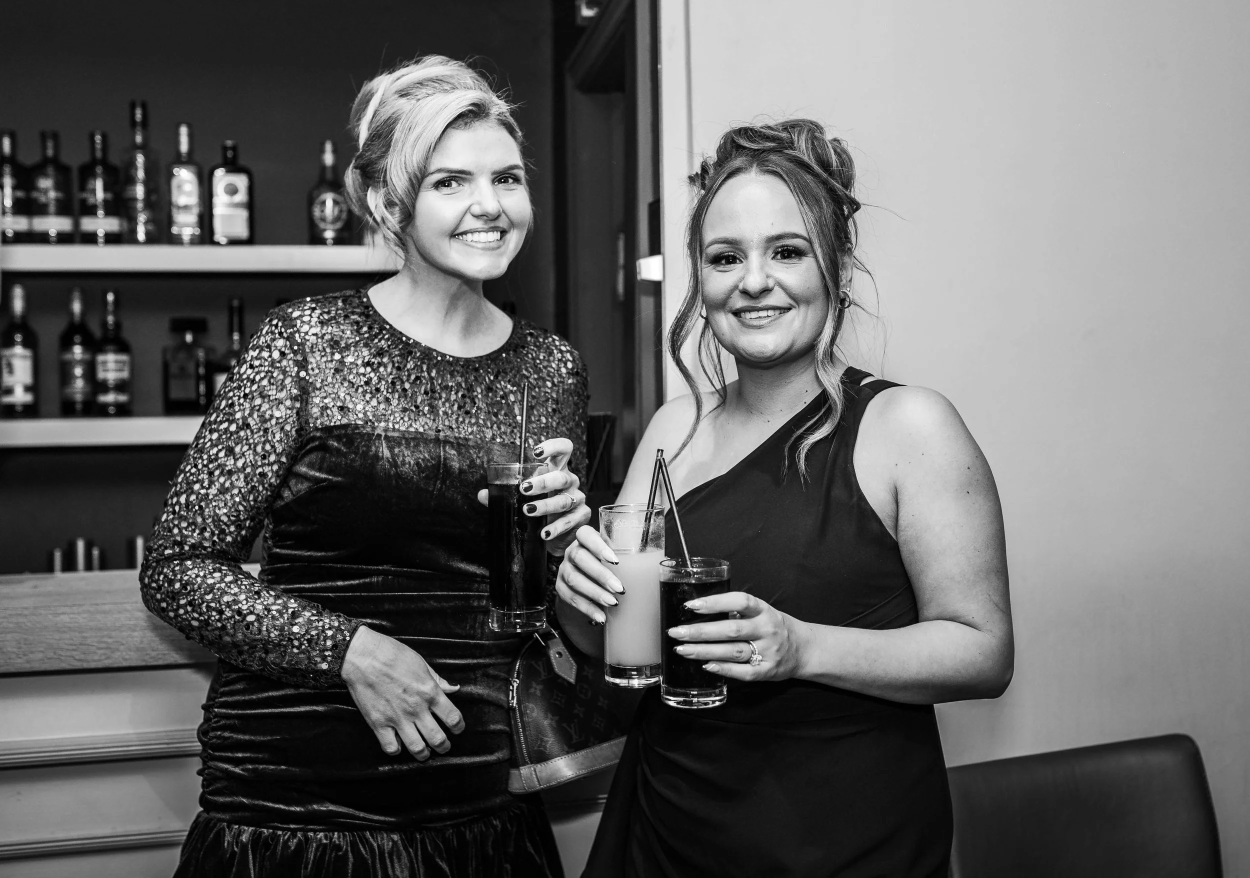 Two women in elegant dresses smiling and holding drinks in a bar or restaurant setting, with bottles on shelves in the background.