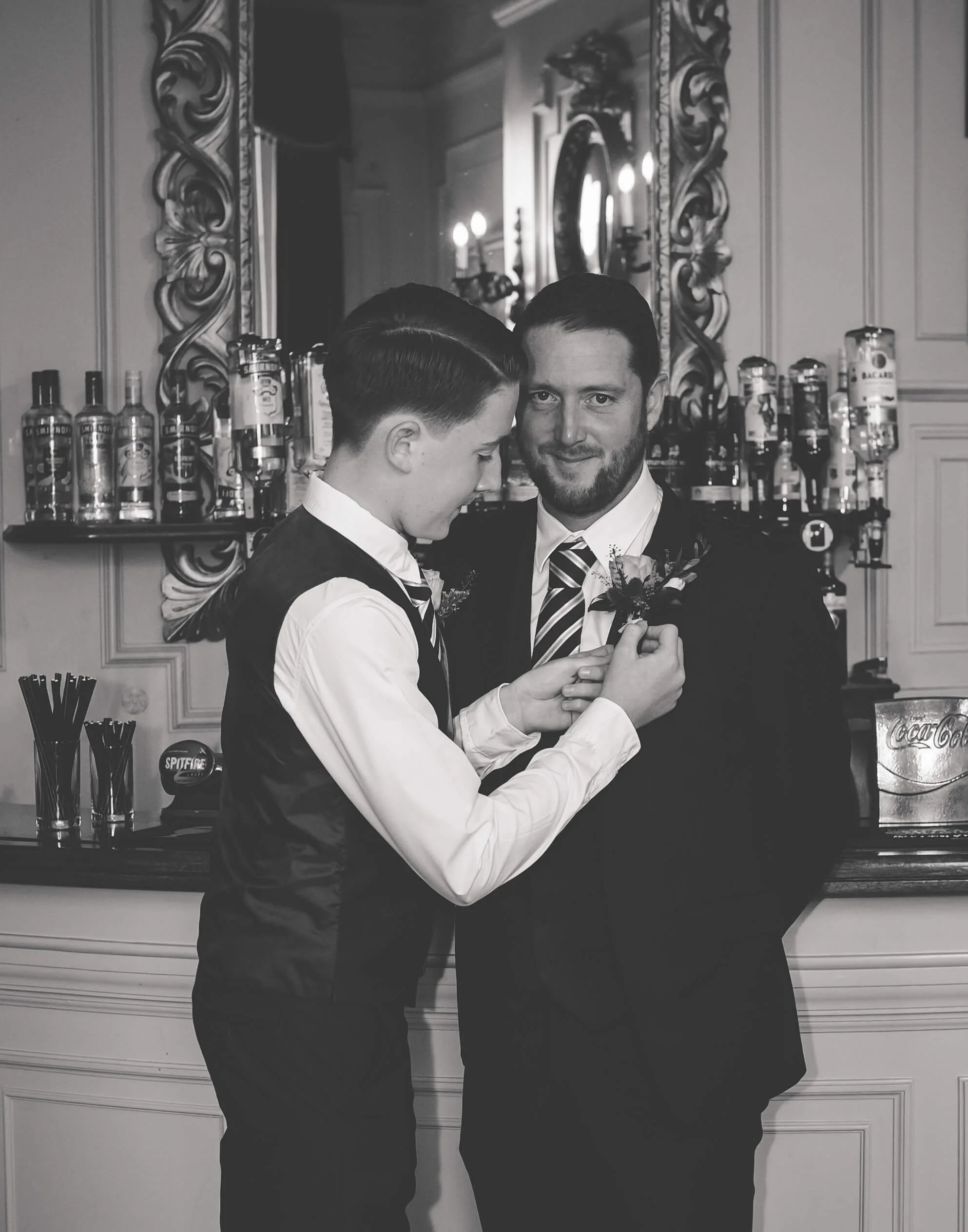 A young man is pinning a boutonniere onto the lapel of an older man, possibly at a wedding or formal event, with a bar and ornate mirror in the background.