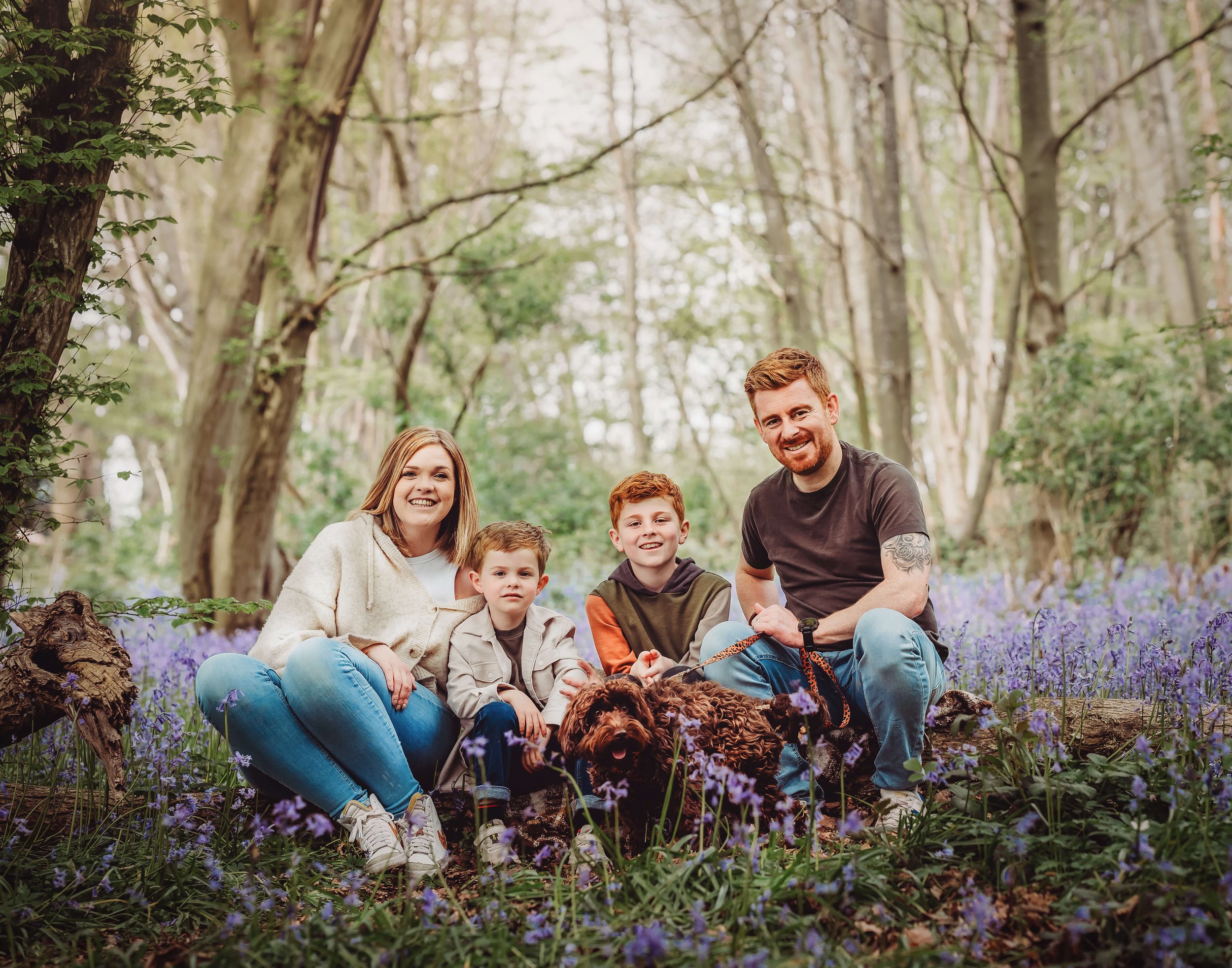 A family of five with a dog sitting on purple flowers in a wooded forest. The family includes a woman, a man, and three boys, all smiling at the camera.