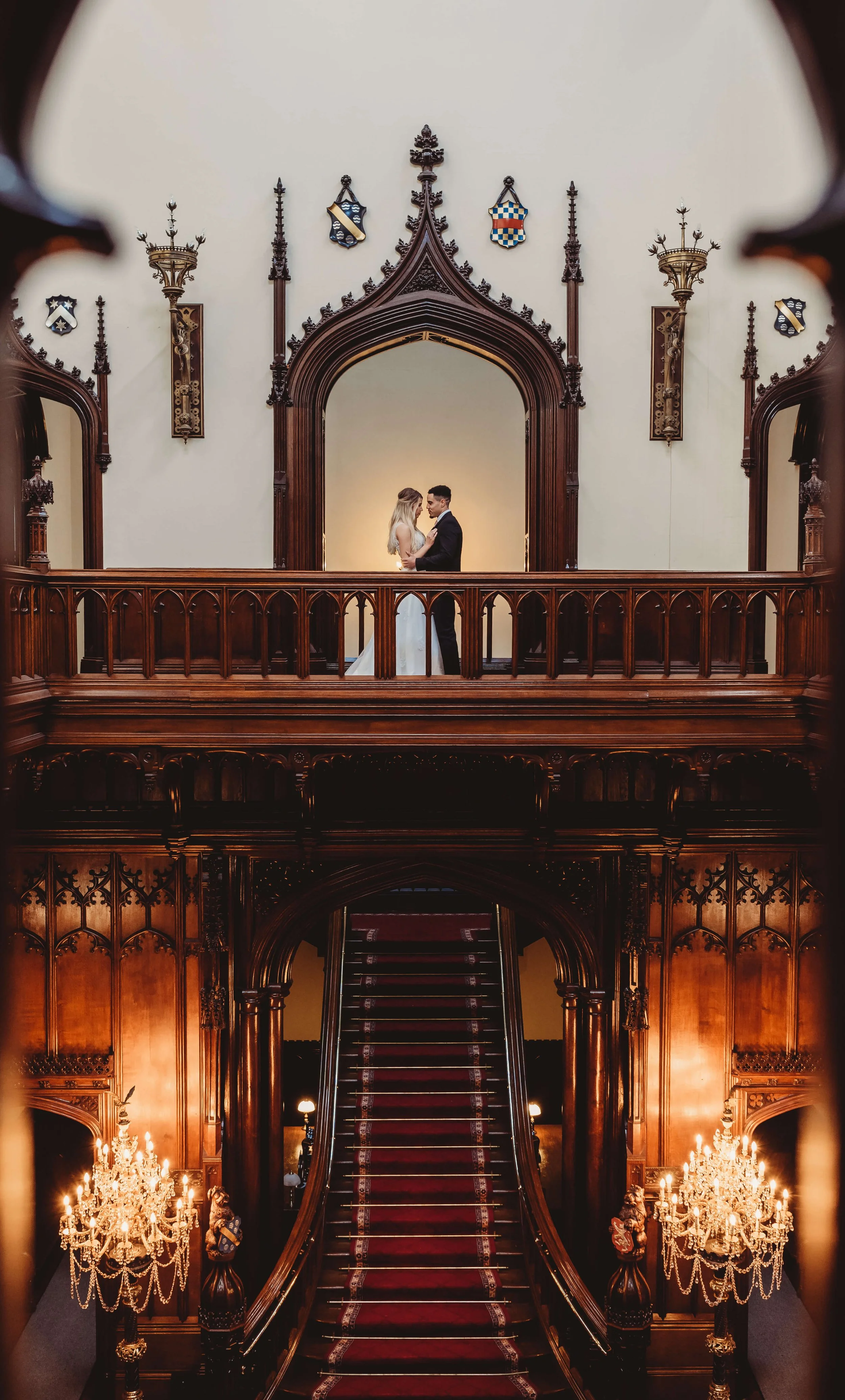 A bride and groom stand in a historic, ornate, wood-paneled hall with chandeliers, a staircase, and Gothic architectural details.