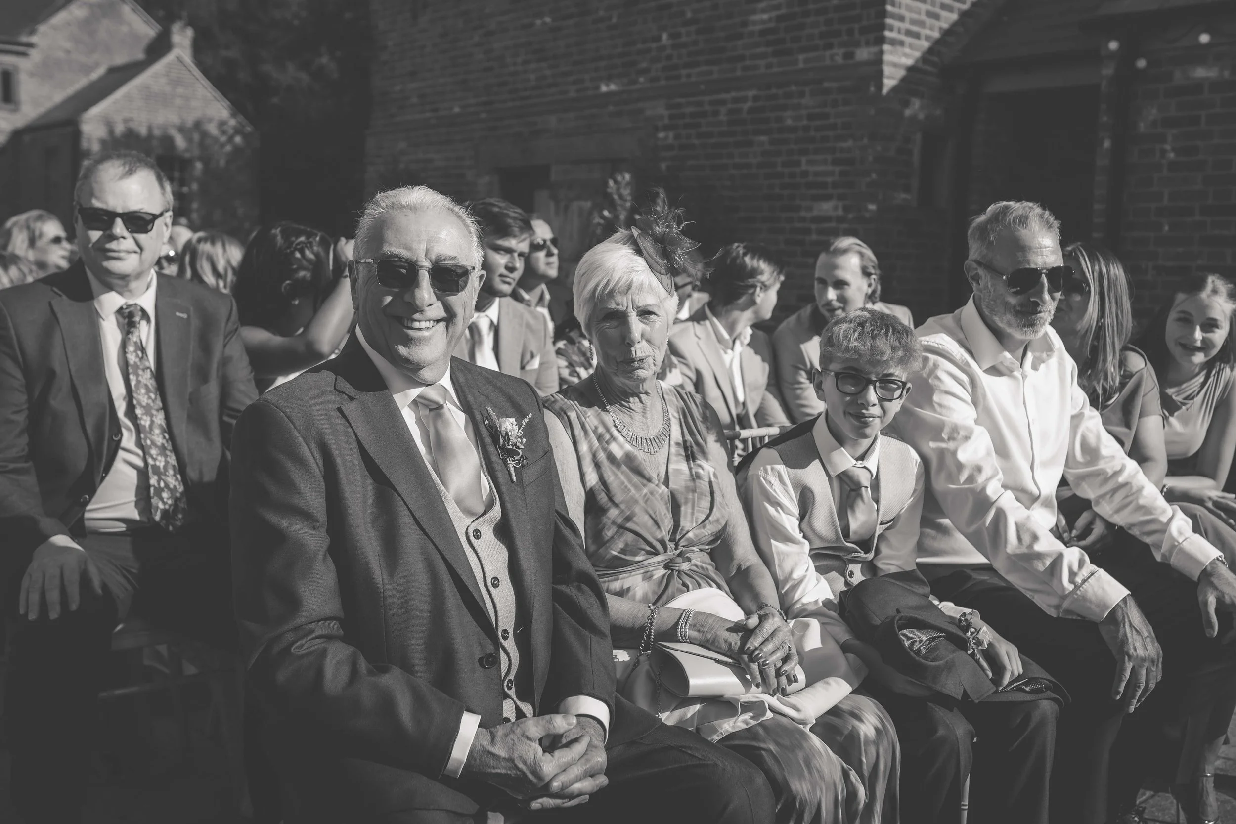A group of people sitting outdoors, dressed in formal attire, attending an event. The photo is in black and white.