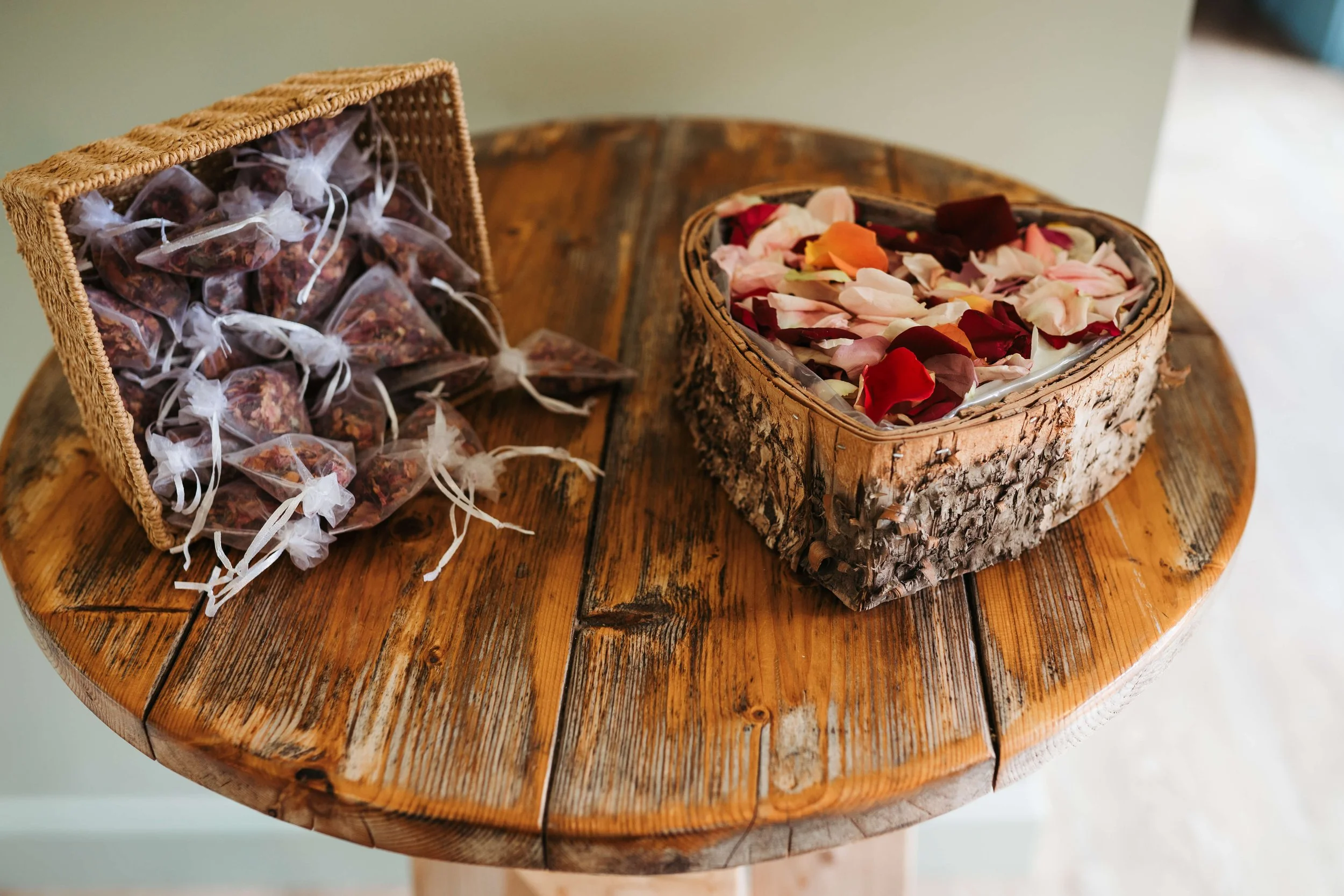A wooden table holds a wicker basket filled with dried flower petals and a glass container with rose petals inside.