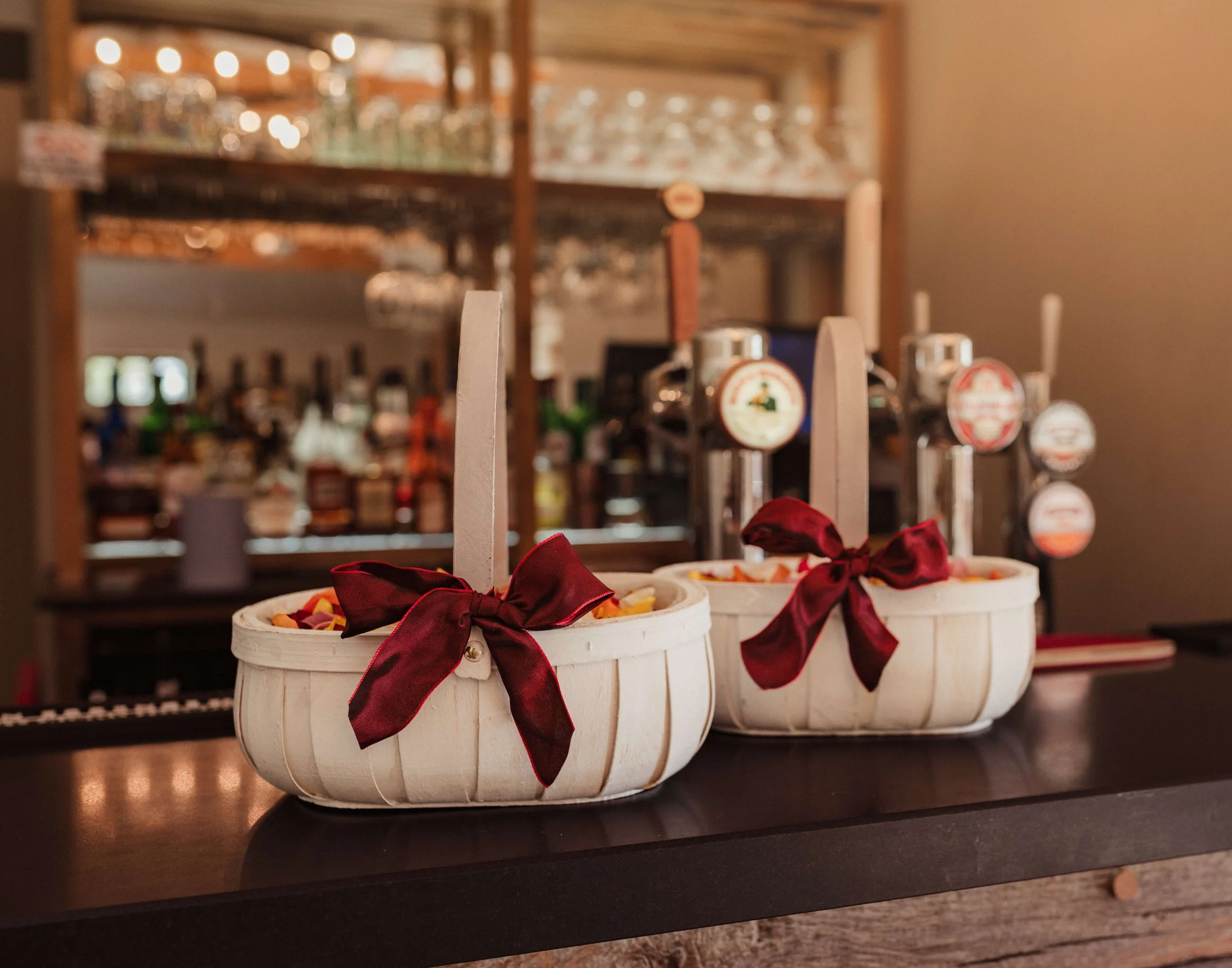 Two white baskets with red ribbons, filled with candy, placed on a dark wooden bar counter in a restaurant or bar setting.