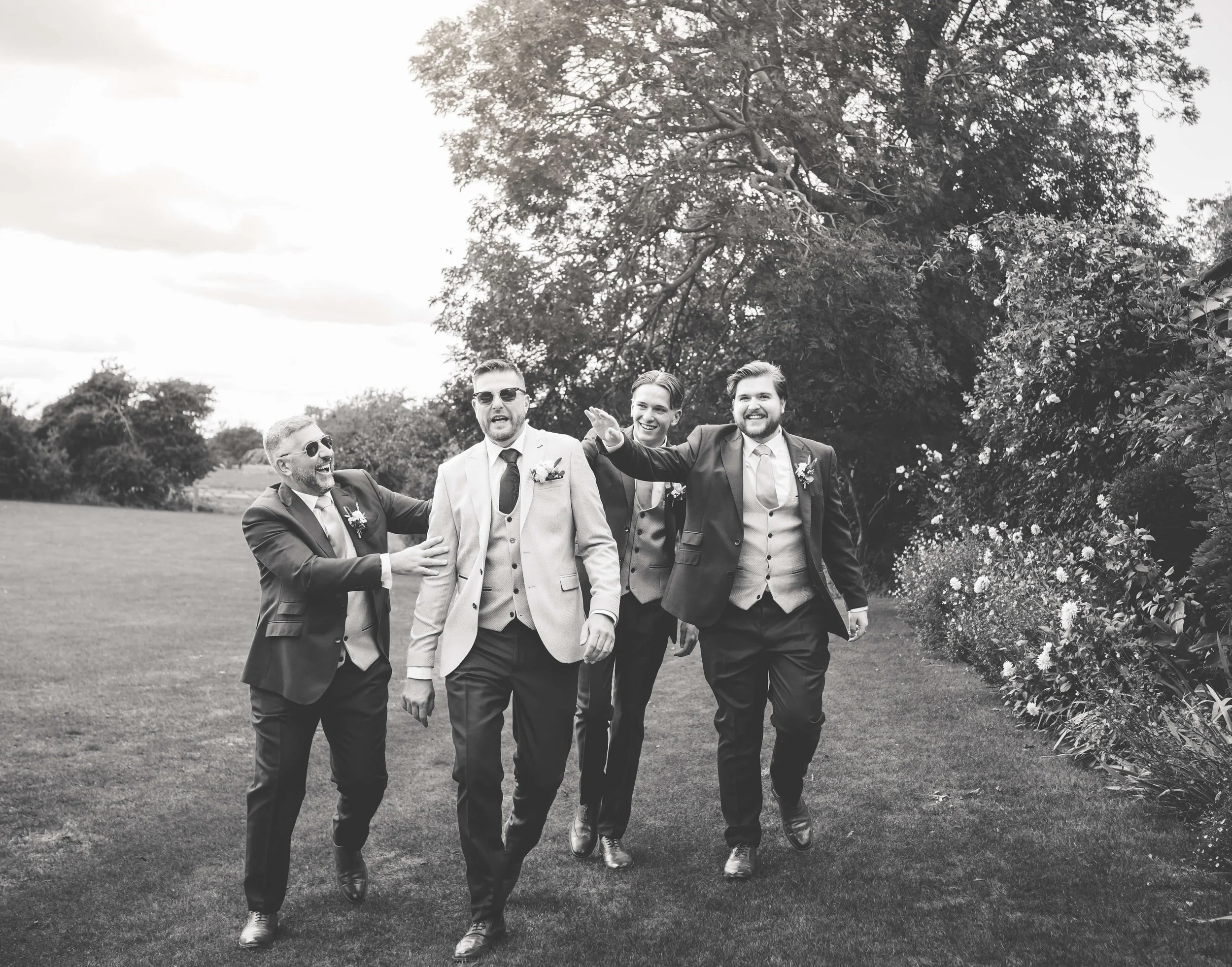 Group of five men walking outdoors dressed in formal suits, smiling and enjoying each other's company, with trees and shrubs in the background.
