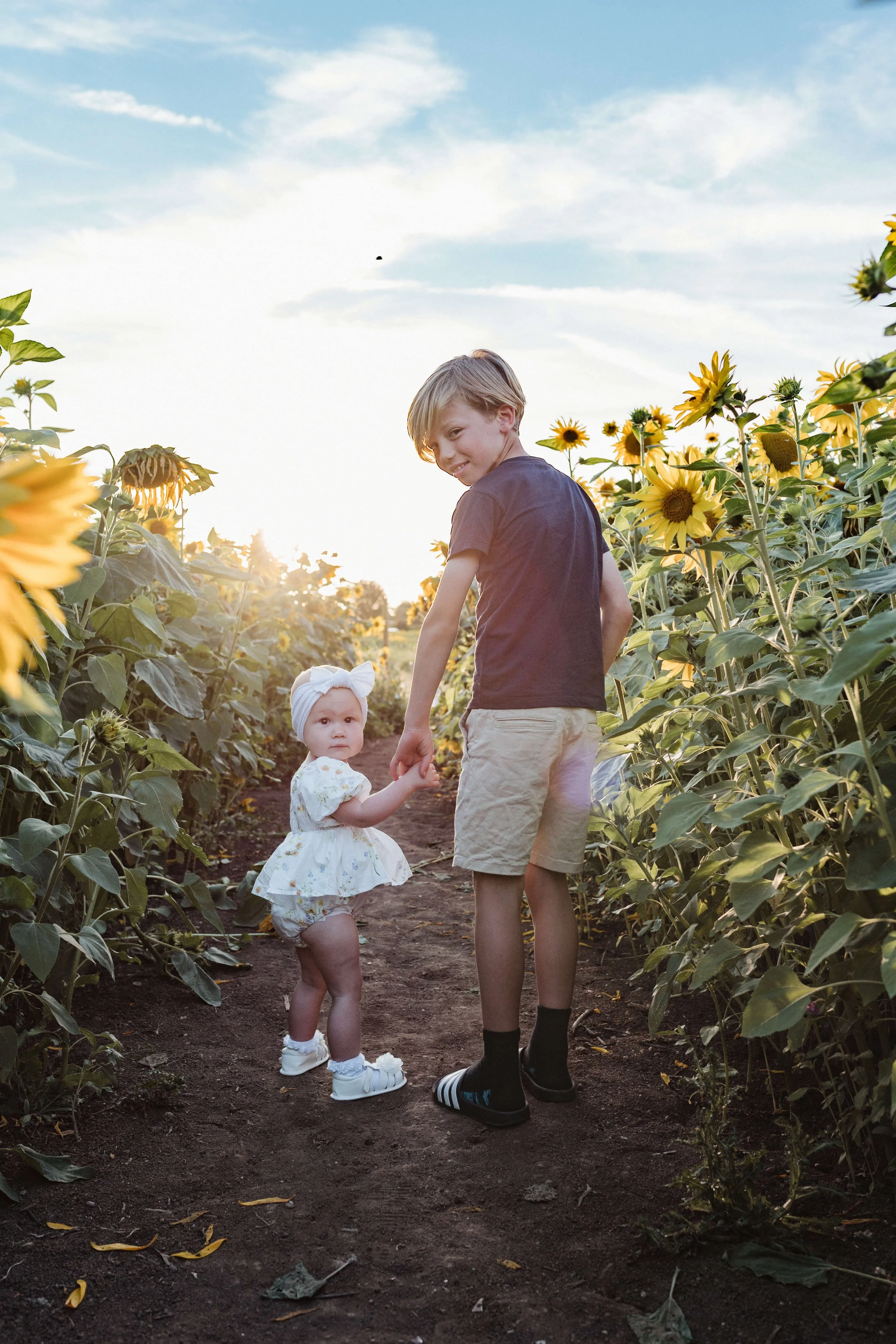 A boy and a baby girl holding hands in a sunflower field during sunset, with blue sky and clouds in the background.