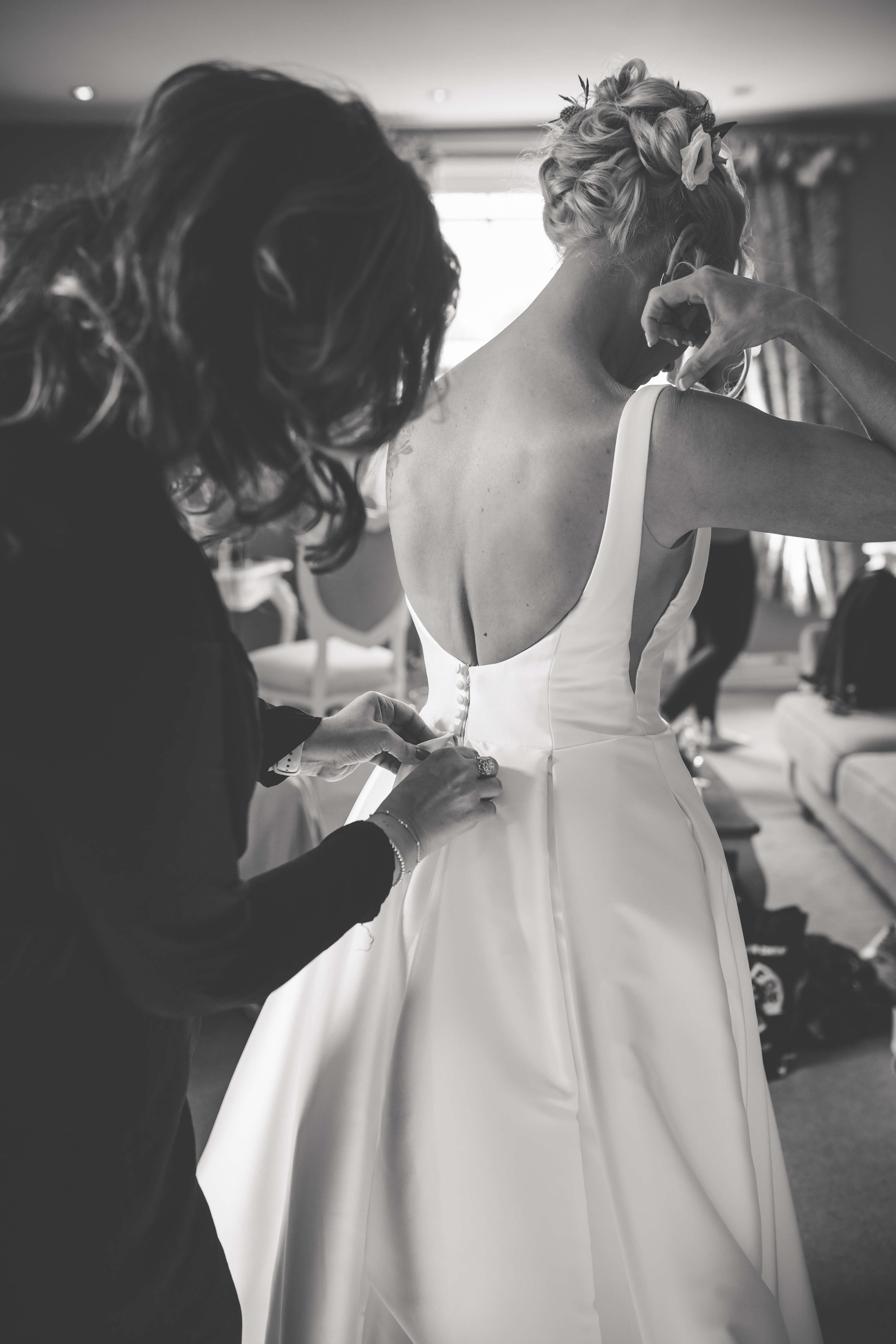 A bride in a wedding dress being helped with her dress by a woman in a black outfit, as she gets ready for her wedding in a room with curtains and furniture.