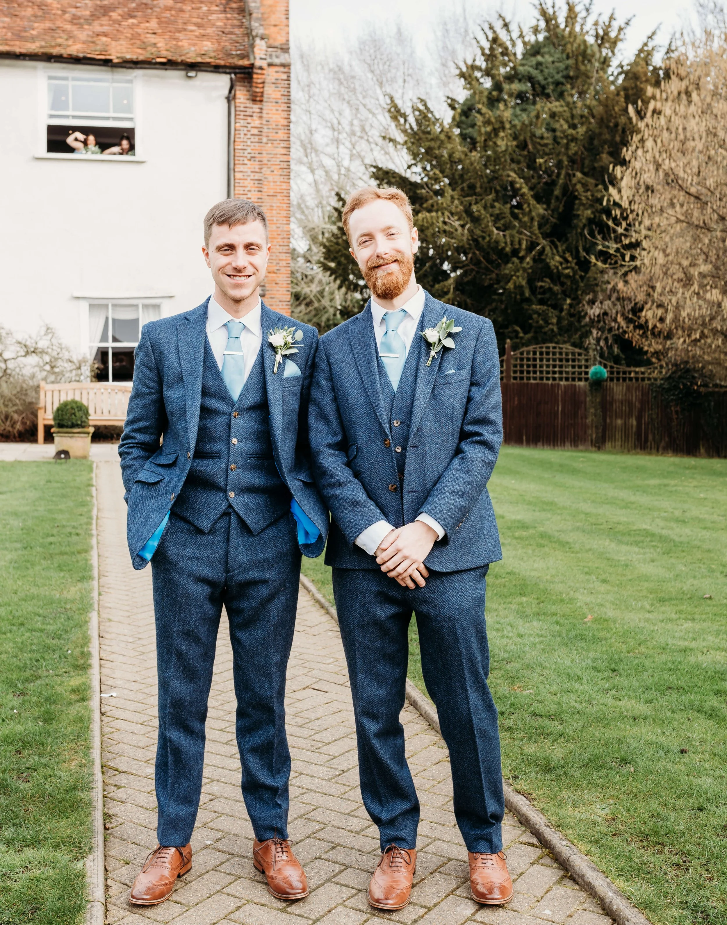 Two men in matching blue suits and ties, with white shirts and boutonnières, standing on a stone pathway in a grassy outdoor setting, smiling at the camera.