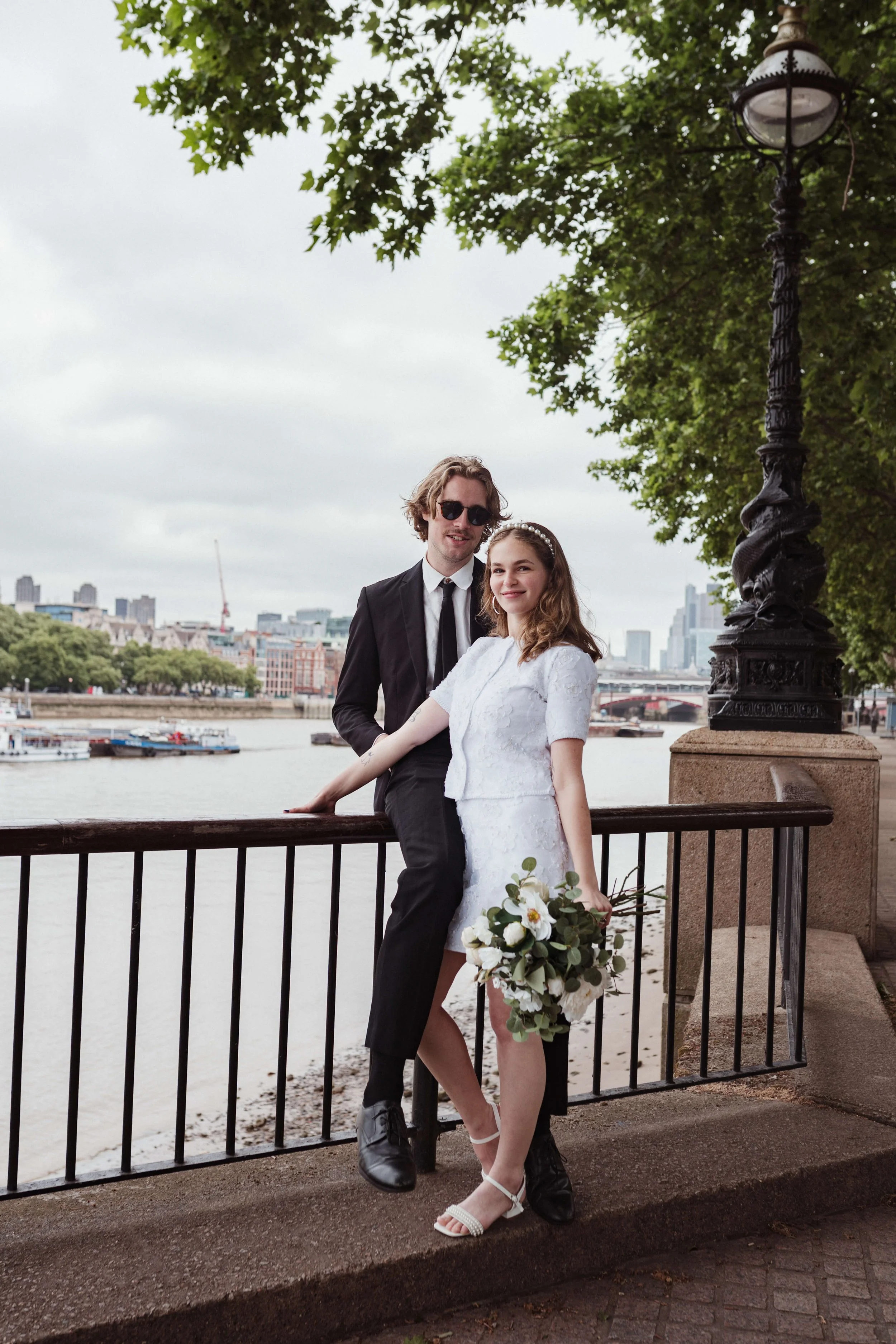 A couple dressed in wedding attire standing by a river on a cloudy day, with city buildings in the background. The woman is holding a bouquet of flowers, and the man is wearing sunglasses.
