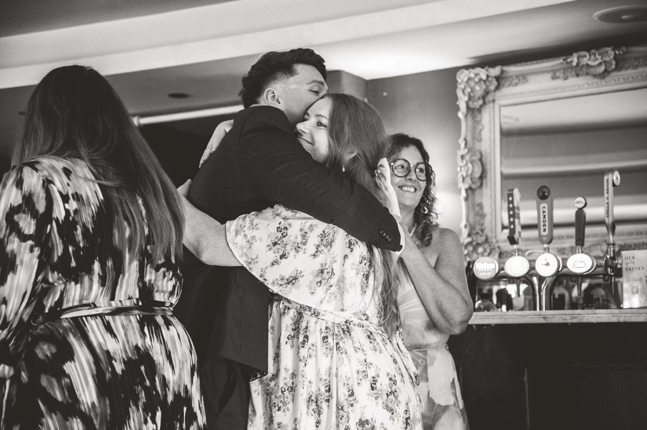 Four women sharing a joyful hug and smiling in a bar or restaurant setting with a large ornate mirror and beer taps in the background.