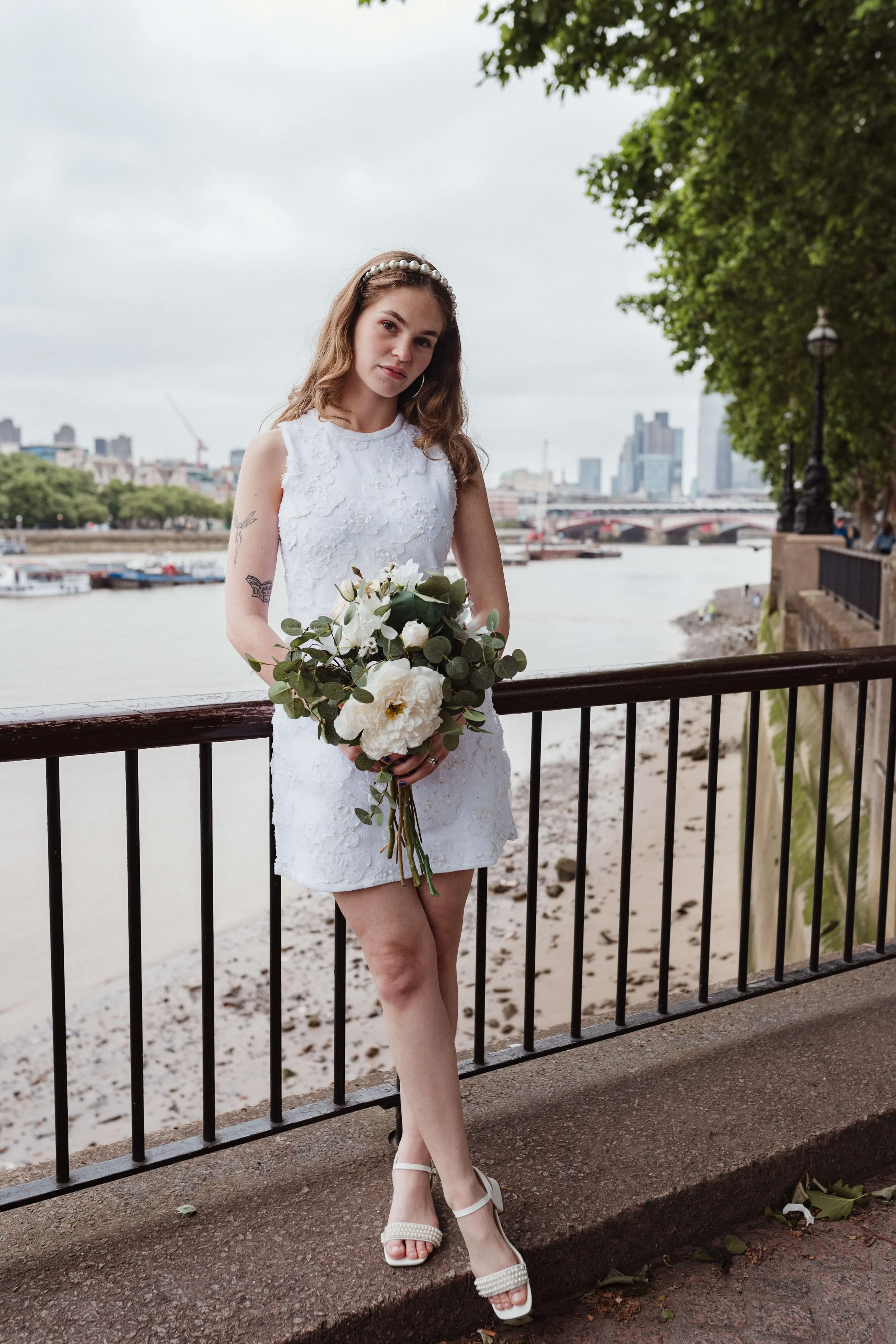 A young woman in a white lace dress standing by a riverside railing, holding a bouquet of white flowers and greenery, with a city skyline in the background.