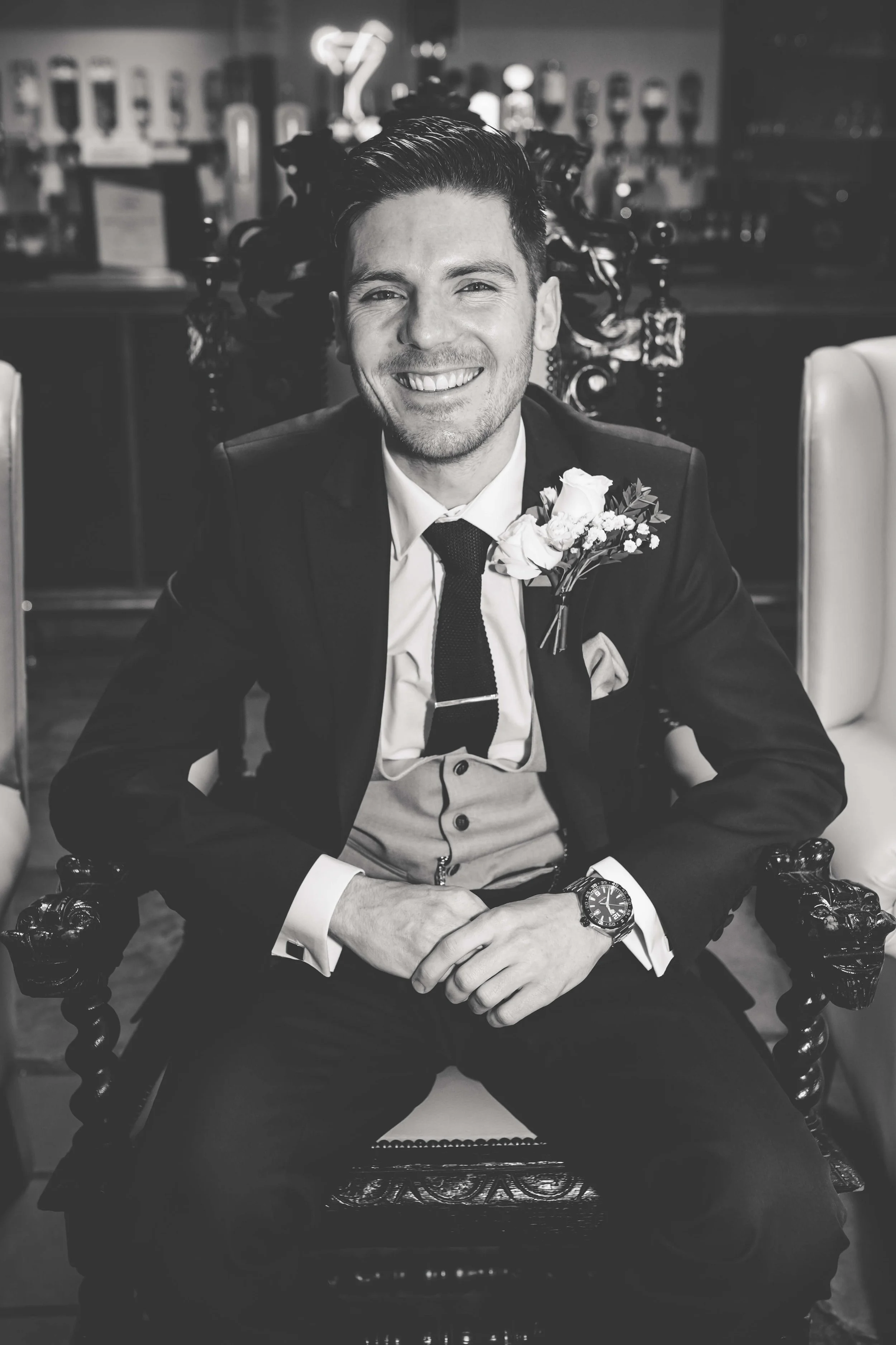 A man in a suit sitting in an ornate chair, smiling, with a boutonniere on his lapel, in a bar or restaurant setting.