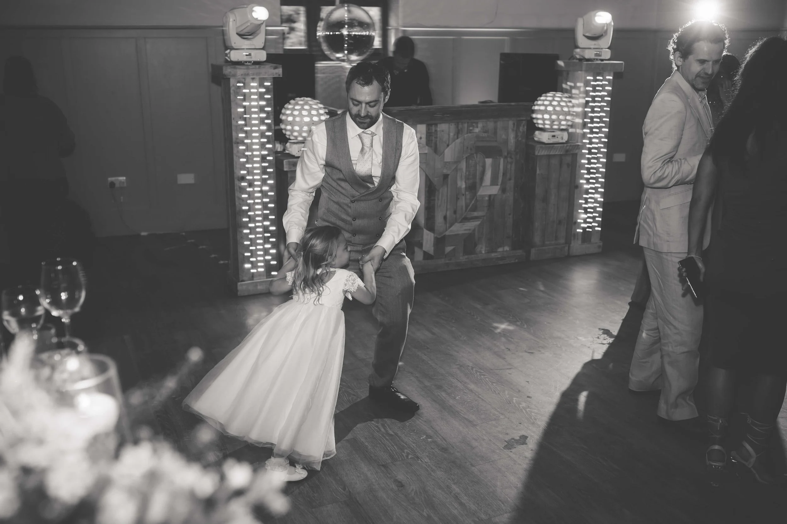 A man in formal attire dancing with a young girl in a white dress at a party or event, with other people in the background and a decorated DJ booth.