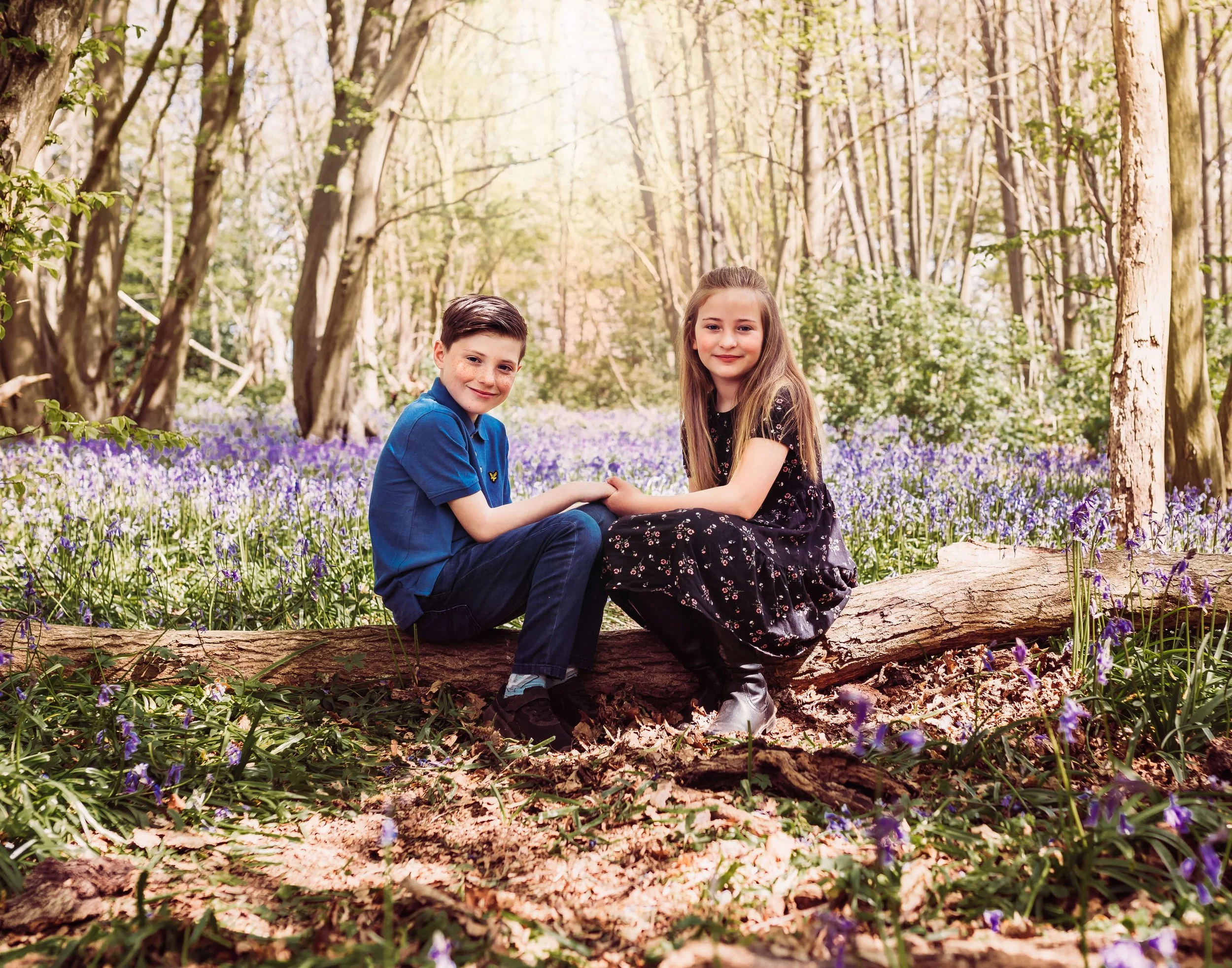 A young boy and girl sitting on a fallen tree trunk in a forest clearing filled with purple flowers and green trees, holding hands and smiling at the camera.