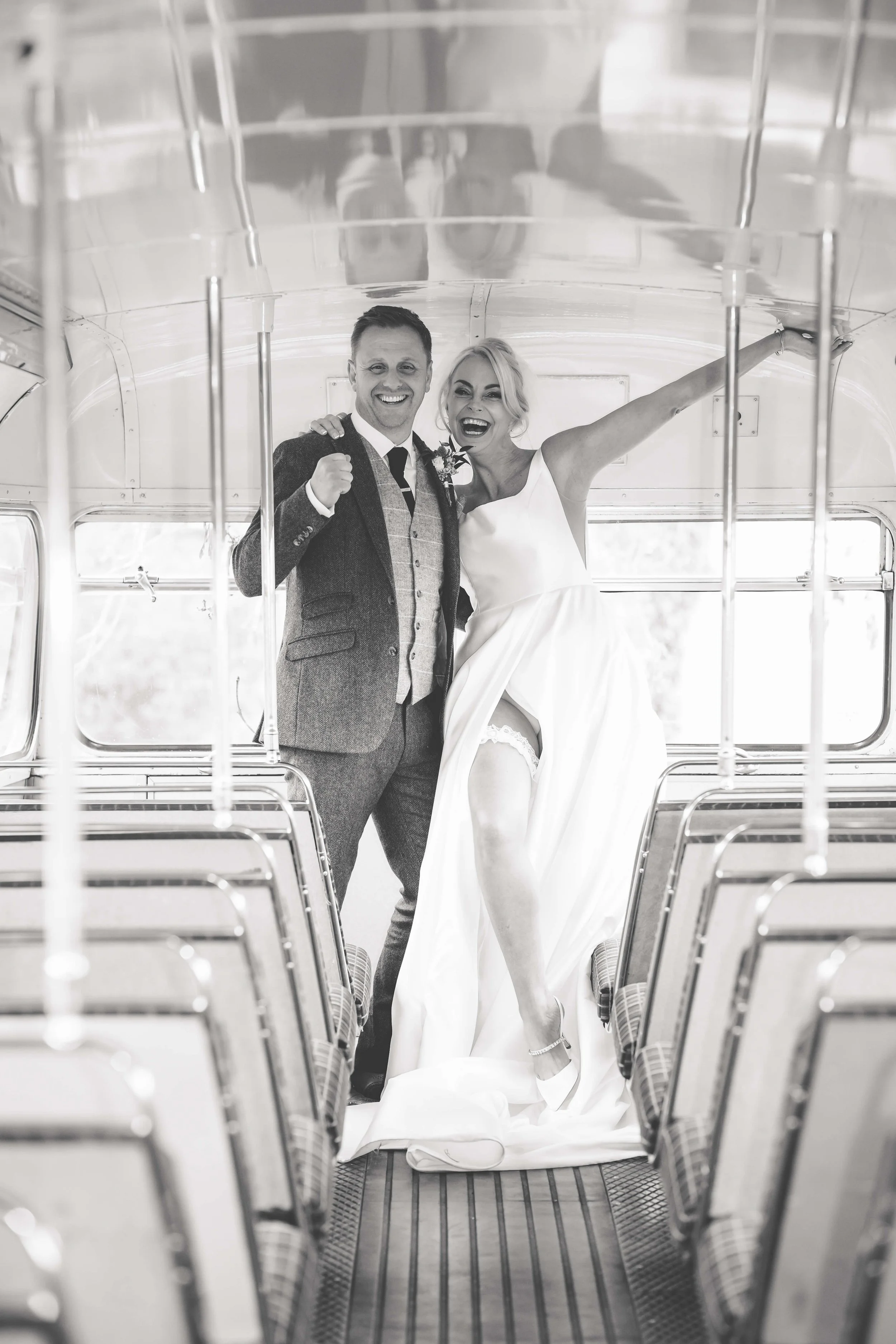 Black and white photo of a smiling man in a suit and a woman in a wedding dress, both standing inside an empty bus, celebrating and showing joy.