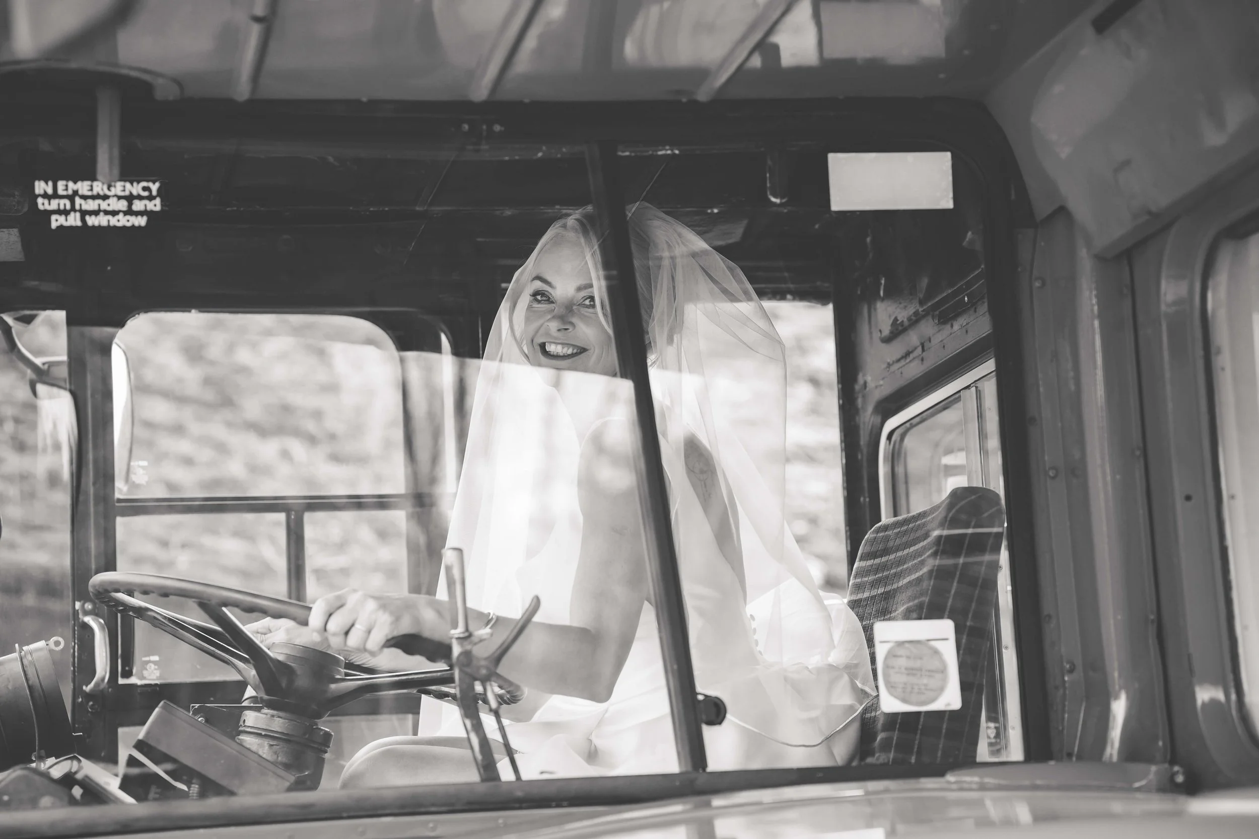 A woman in a wedding dress with a veil, smiling while sitting in the driver's seat of a bus.