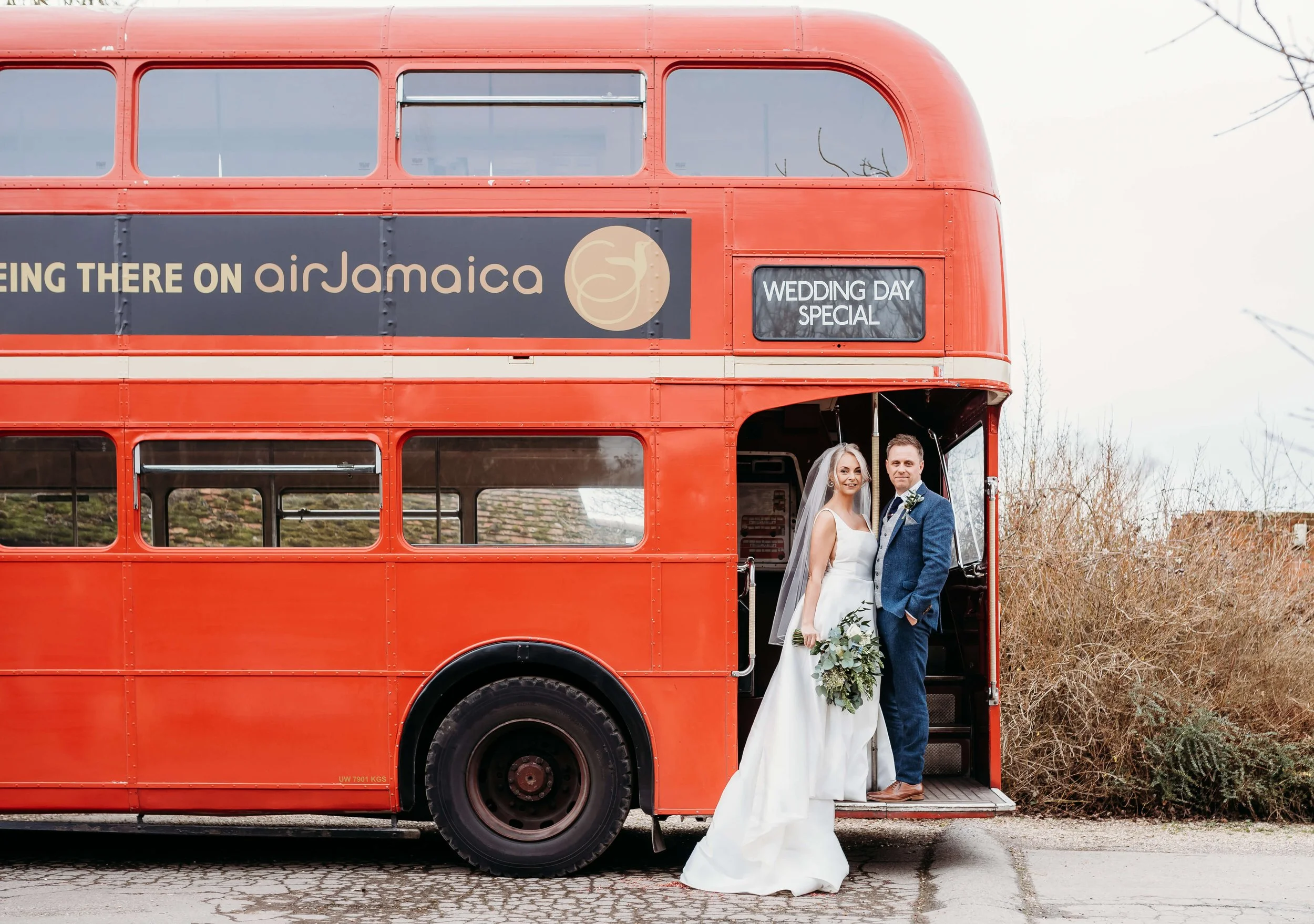 A bride and groom standing on the steps of a vintage red double-decker bus, with the bride holding a bouquet of greenery and the groom in a blue suit. The bus displays the text 'Flying There on airJamaica' and 'Wedding Day Special'.