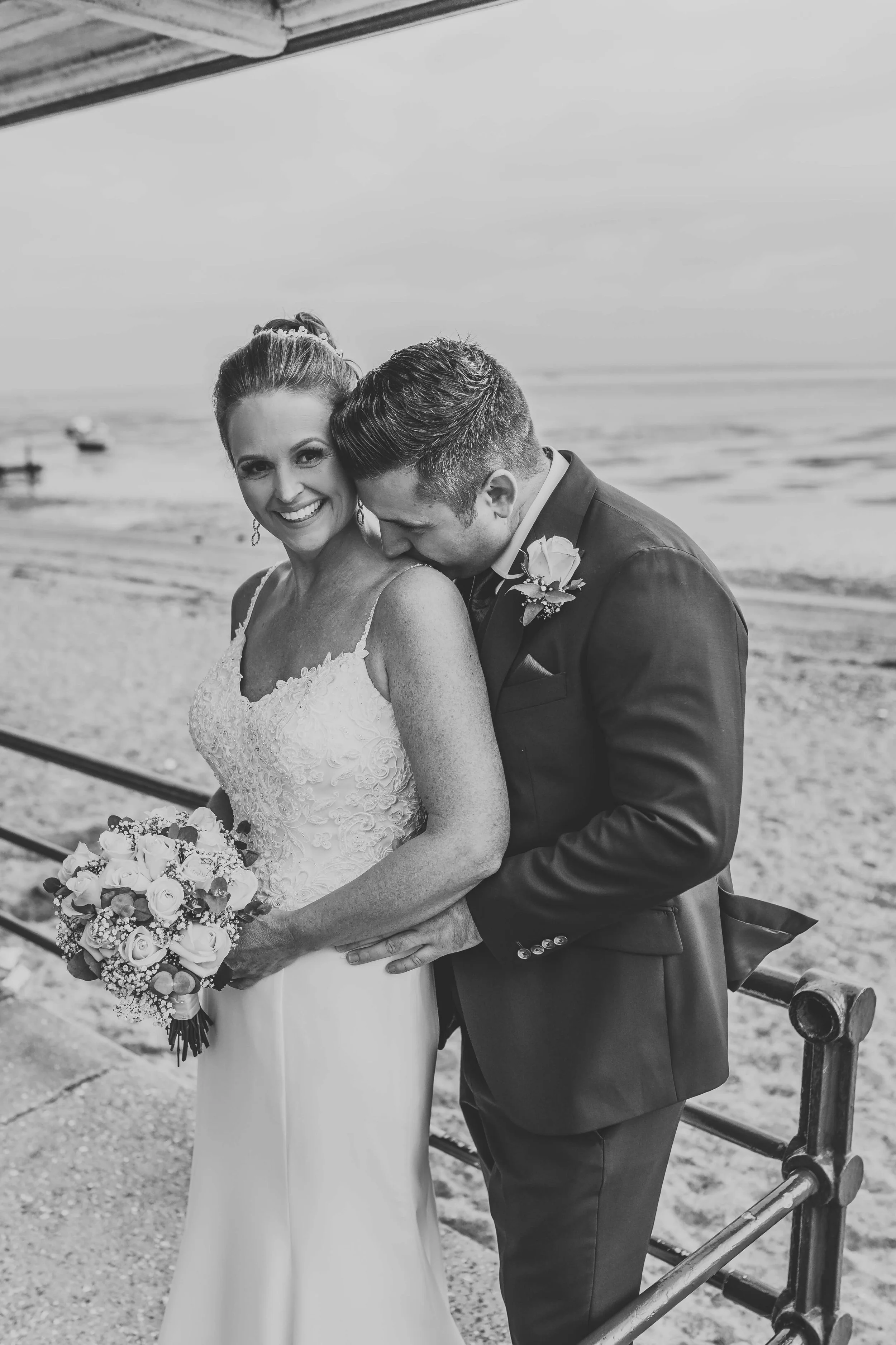 Black and white photo of a bride and groom at the beach, the groom is kissing the bride's shoulder while she smiles at the camera, holding a bouquet of flowers.