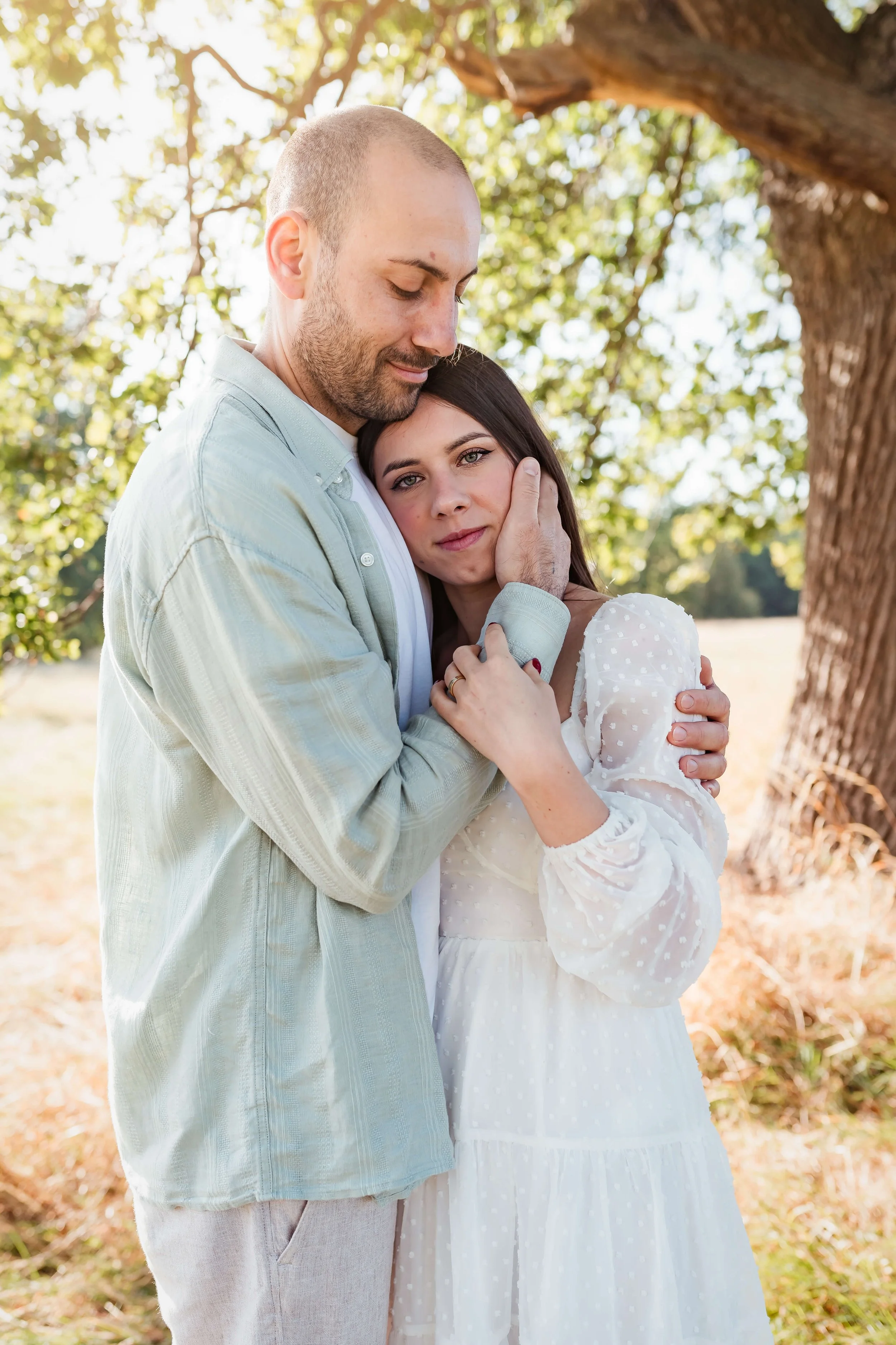 A couple embracing outdoors beneath a large tree with sunlight filtering through the leaves, the man gently holding the woman's face as they look emotional.