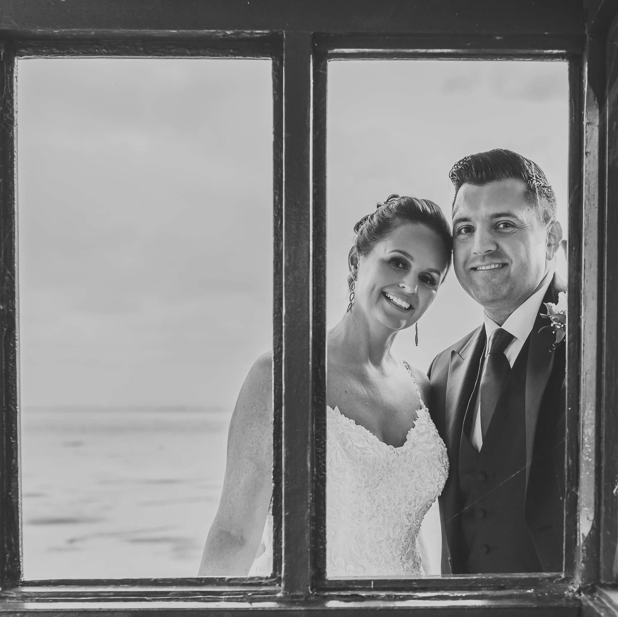 A black and white photograph of a newlywed couple smiling, seen through a window divided into four panes, with a beach and ocean in the background.