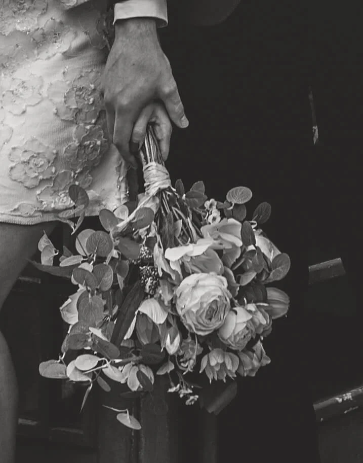 A person holding a bouquet of flowers in their hand, with part of a wedding dress visible nearby.