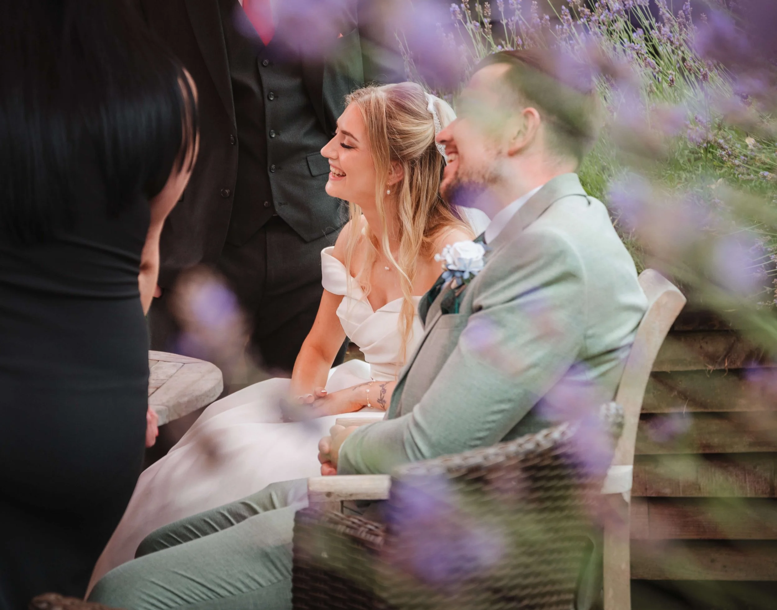 A bride and groom seated outdoors during their wedding ceremony, smiling and laughing, surrounded by purple flowers and a wooden backdrop.