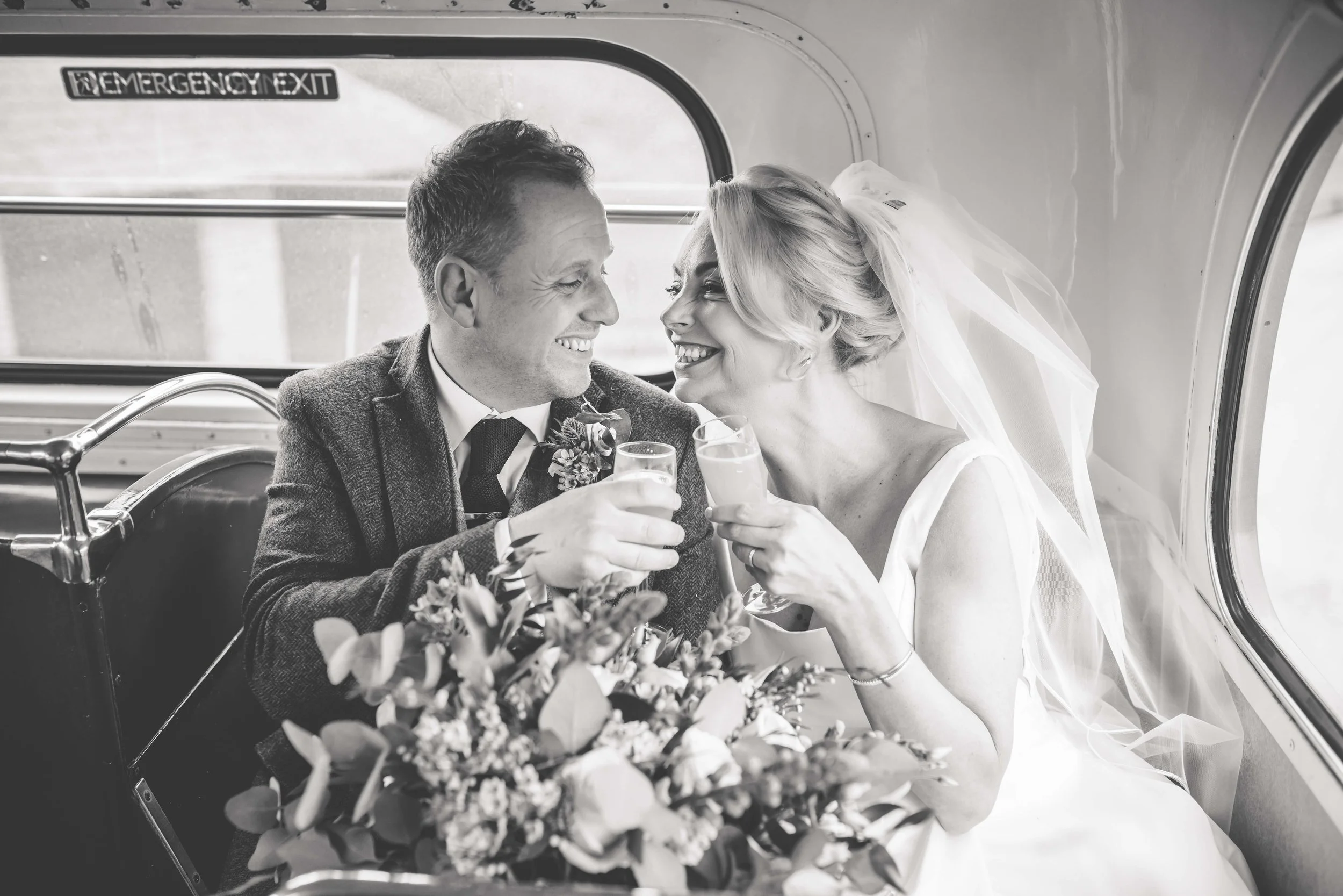 A newlywed couple in wedding attire sitting inside a vehicle, smiling and clinking glasses, with a bouquet of flowers in front of them.