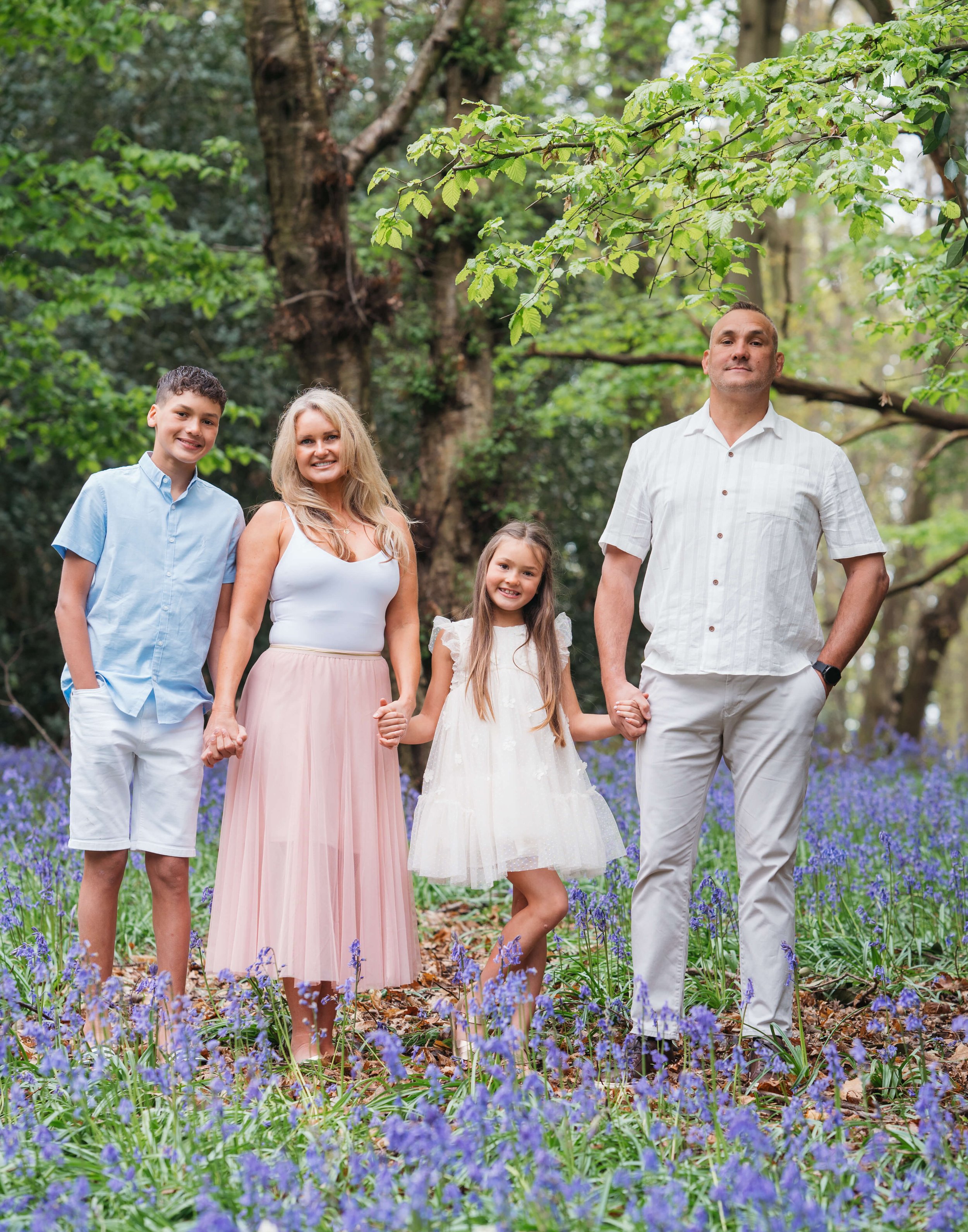 A family of five standing in a forest clearing with purple flowers, holding hands and smiling.