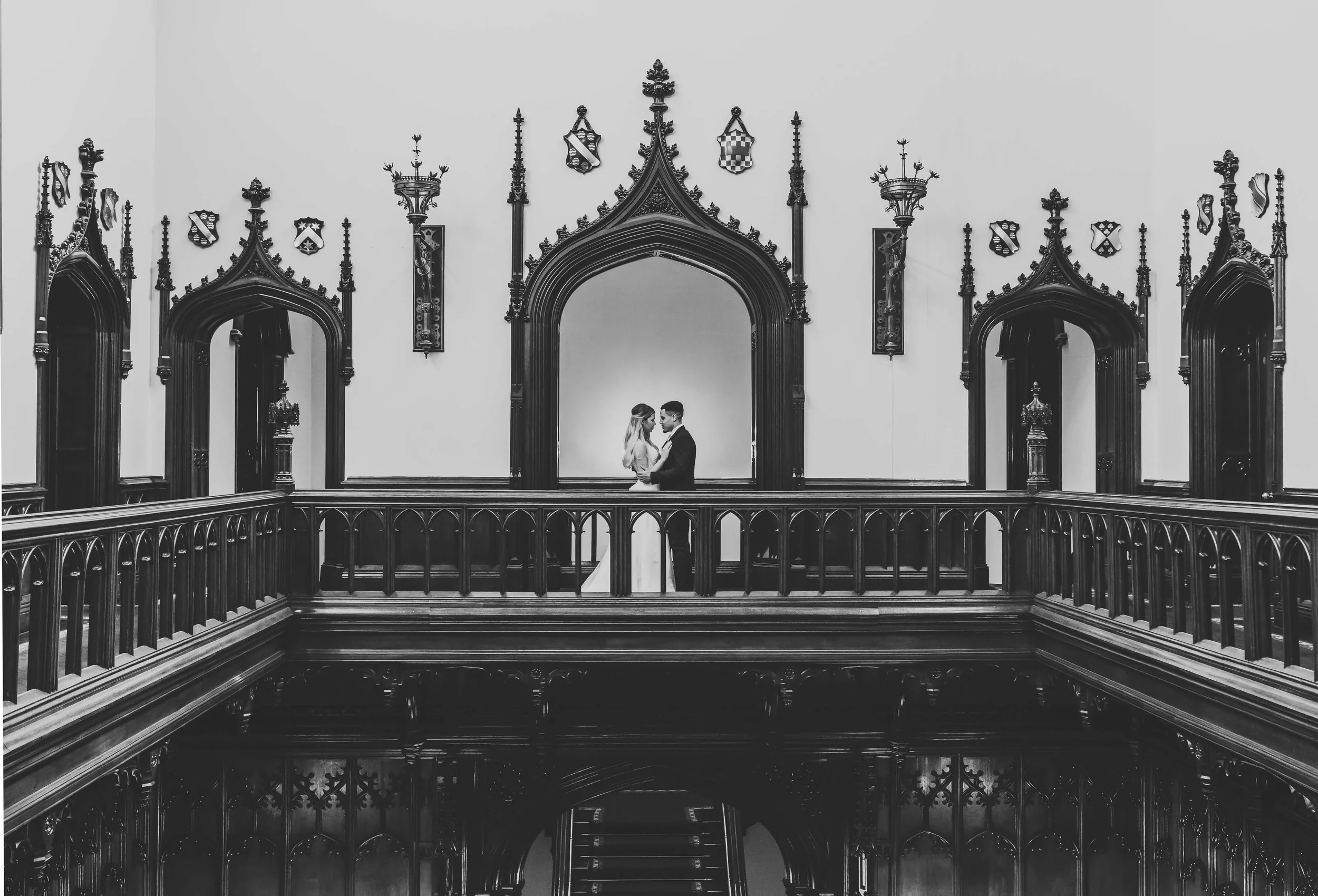 A black and white photo of a couple standing on a balcony inside a historic building with ornate gothic architecture, arched doorways, wood paneling, and decorative shields on the wall.