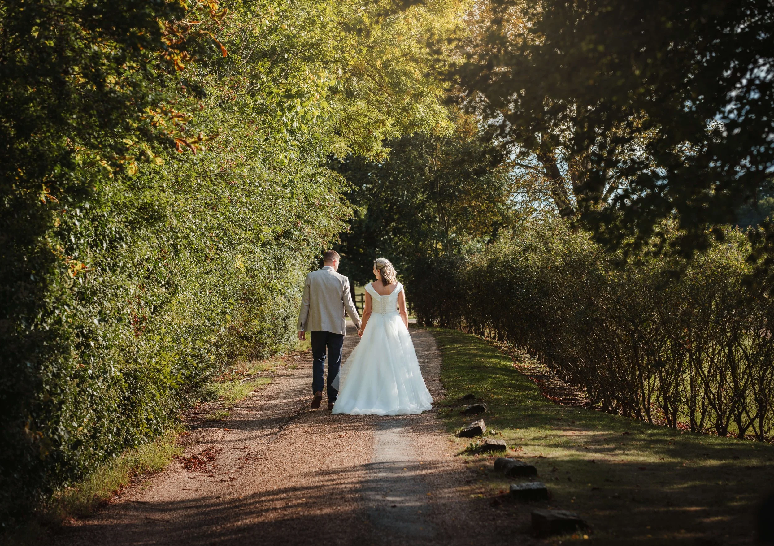 A bride and groom walking hand in hand down a dirt path lined with green trees and bushes, with sunlight filtering through the foliage.