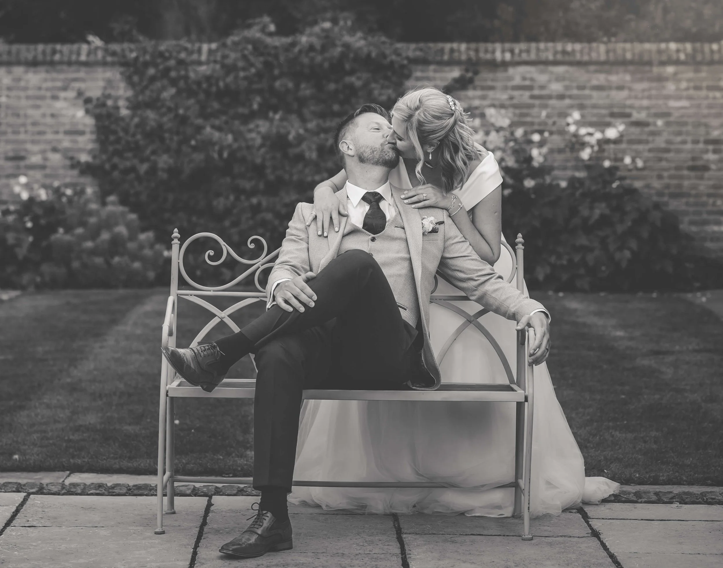 A black-and-white photo of a couple dressed in wedding attire sitting on a vintage iron bench outdoors. The woman is leaning over to kiss the man on the lips, who has his arm around her. They are in front of a garden with a brick wall in the backgrou