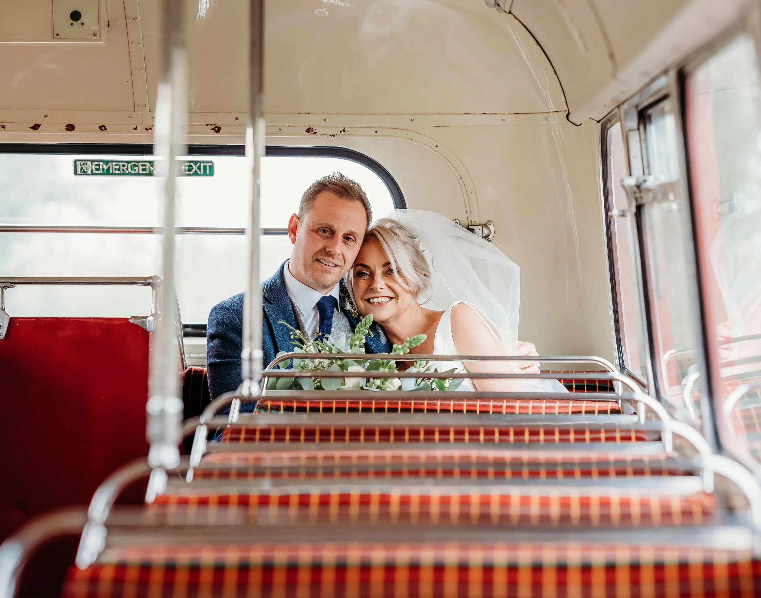 A bride and groom sitting inside a vintage bus, smiling and hugging, with an emergency exit sign visible in the background.