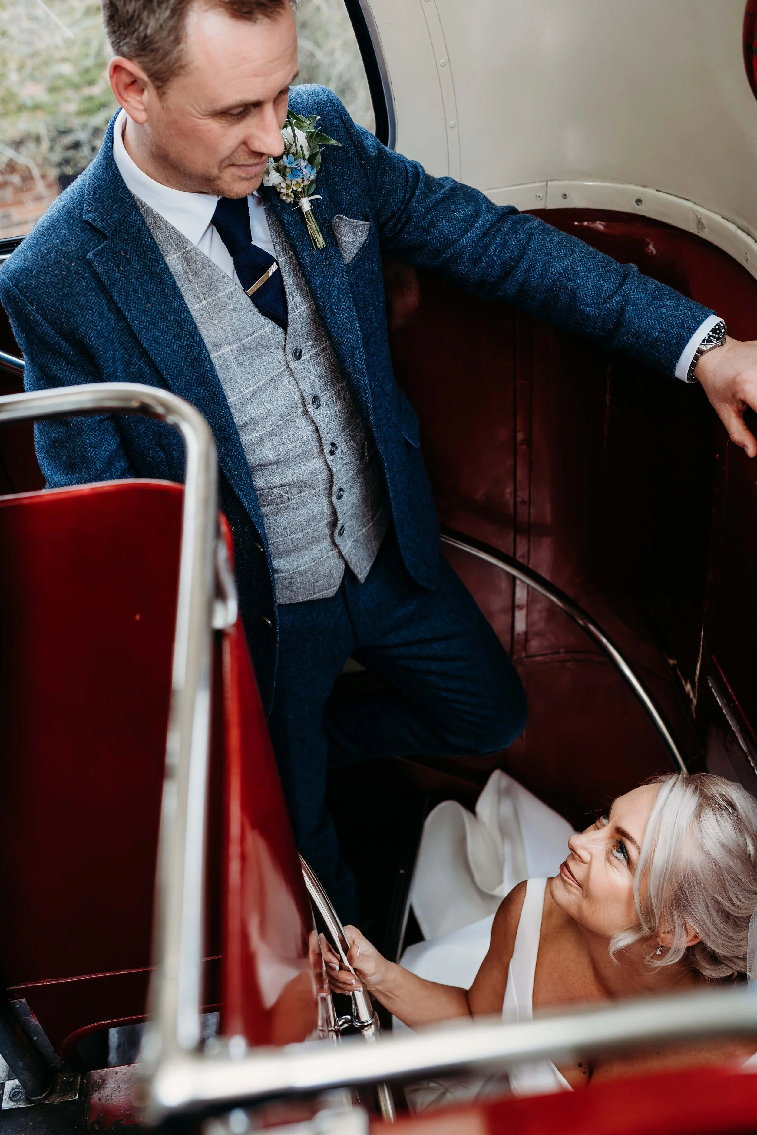 A man in a blue suit and a woman in a white dress inside a vintage red bus, looking at each other, with the woman sitting on the floor and the man standing over her.