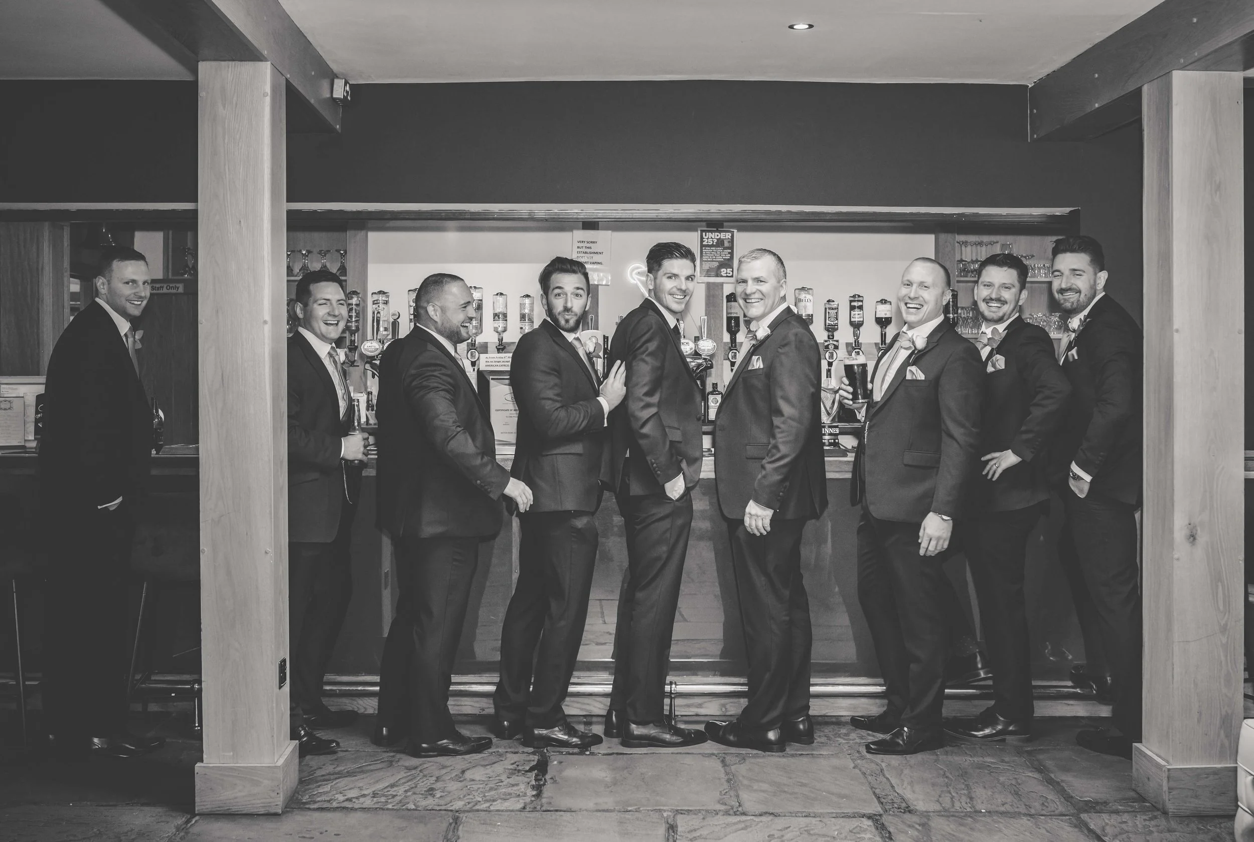 Group of nine men in suits standing at a bar, smiling and posing for a photo.