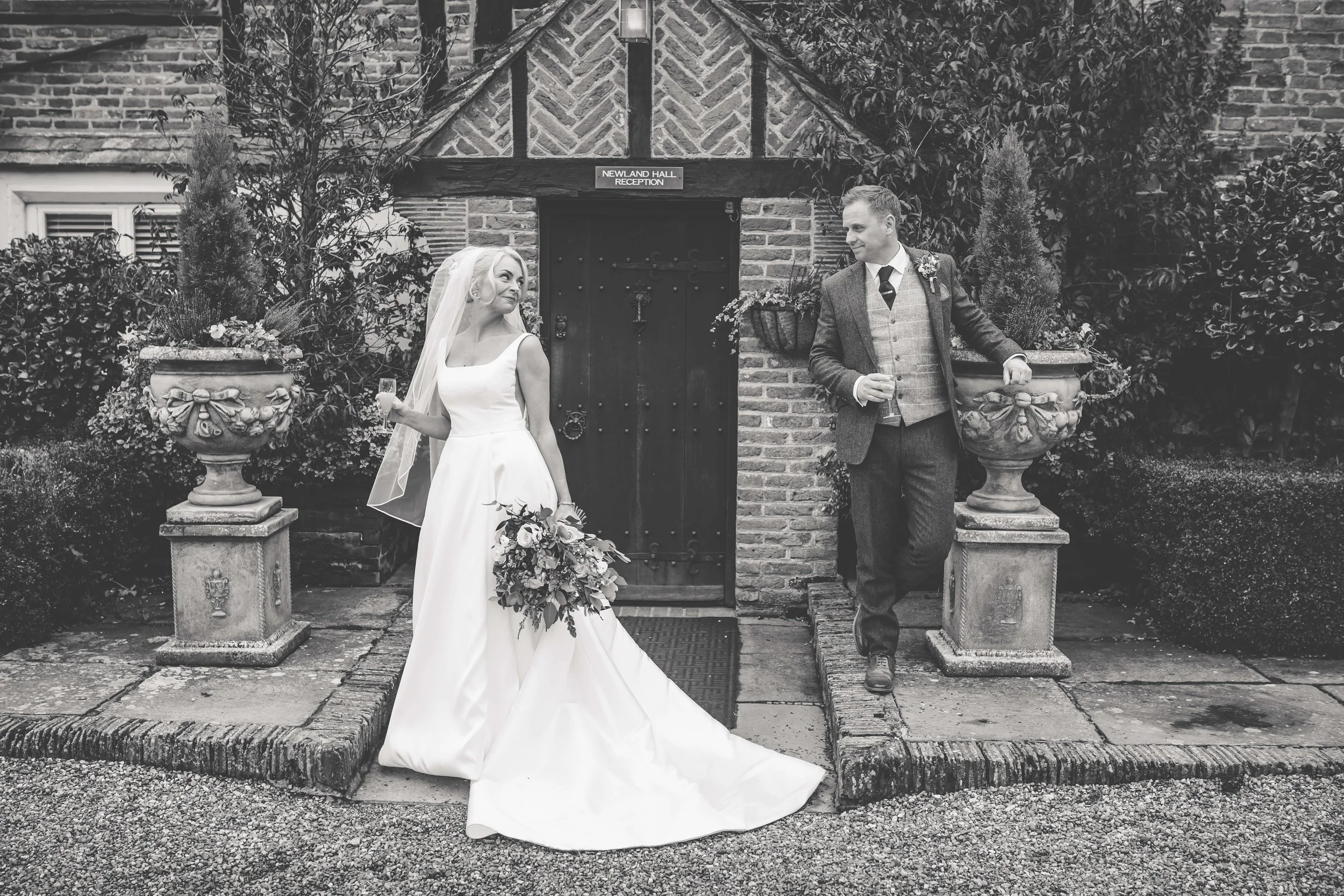 A black and white photo of a bride and groom outside a brick building labeled 'Newland Hall Reception.' The bride is wearing a white wedding dress, holding a bouquet and a glass of champagne, and looking at the groom. The groom is dressed in a suit w