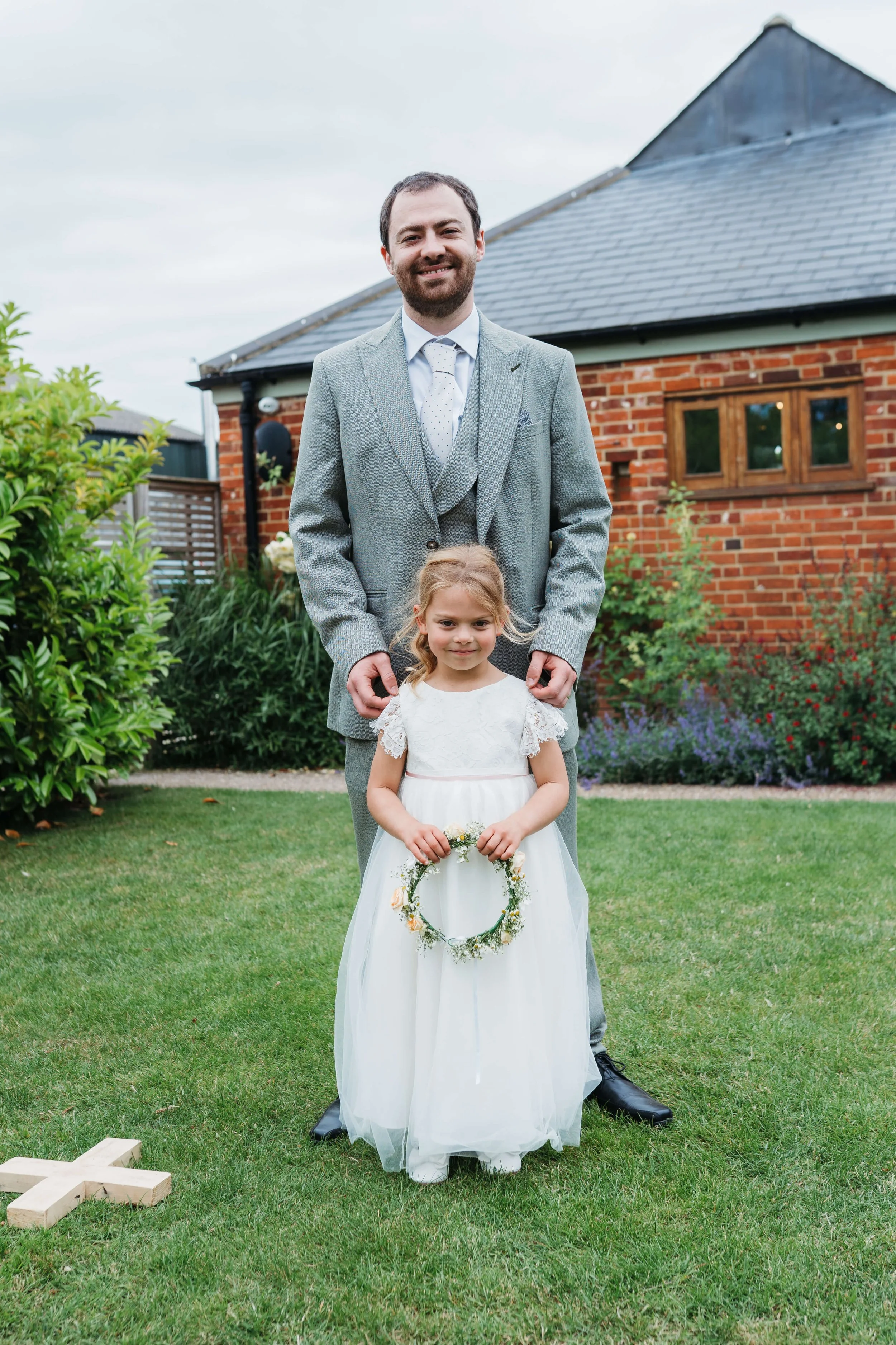 A man and a young girl, possibly a girl, standing outside on a lush green lawn during a wedding ceremony. The girl is dressed in a white dress and holding a floral wreath. The man is dressed in a gray suit and is standing behind her with his hands on